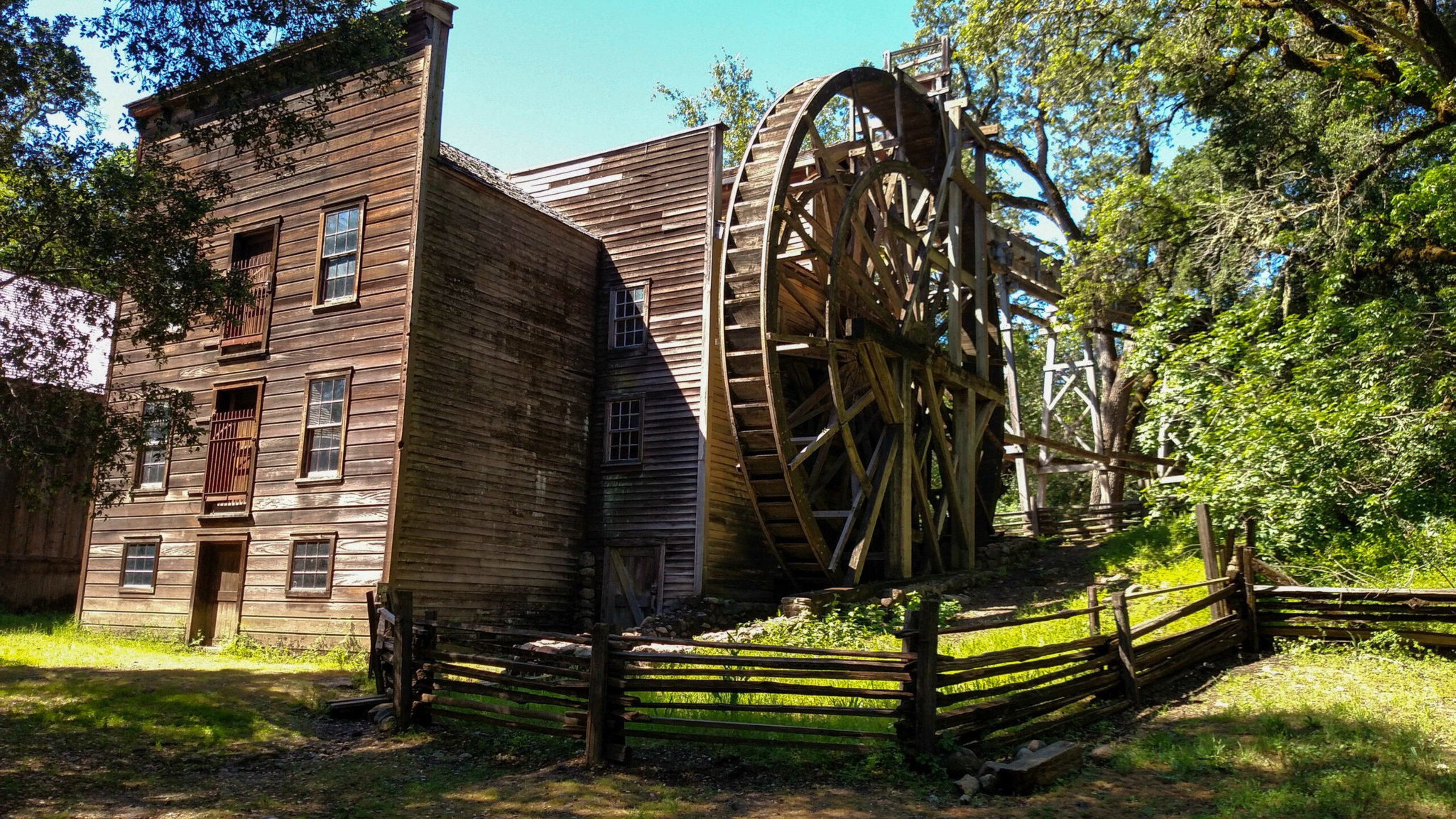 A large wooden watermill with a tall wheel stands beside a rustic building surrounded by a wooden fence and lush green trees.