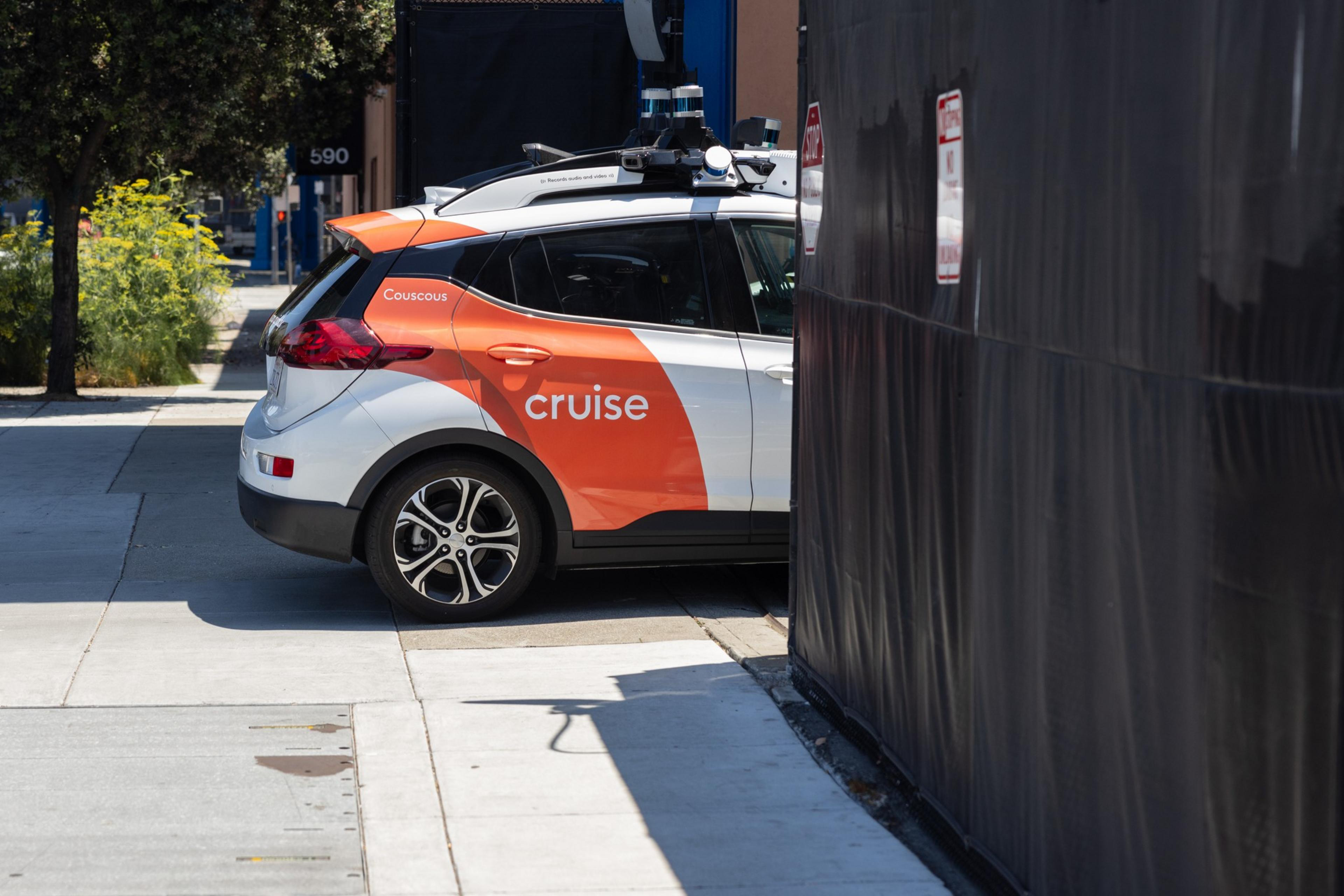 The back half of a white coupe with an orange ribbon on its passenger side showing a Cruise logo pulls into a driveway and enters a fenced-in parking lot.