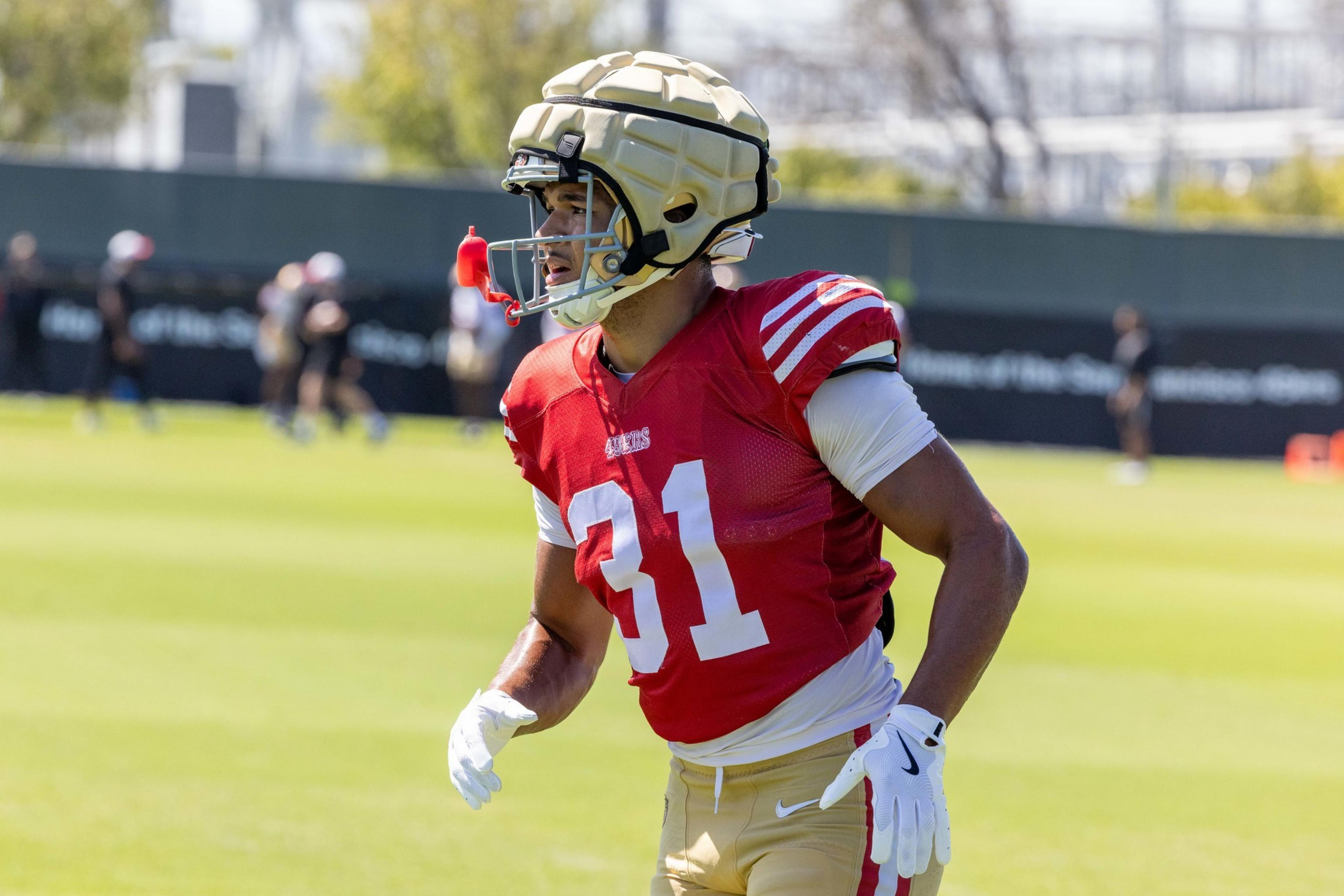 A football player wearing a red practice jersey with number 31, gold pants, a padded helmet, and white gloves is on a grassy field.