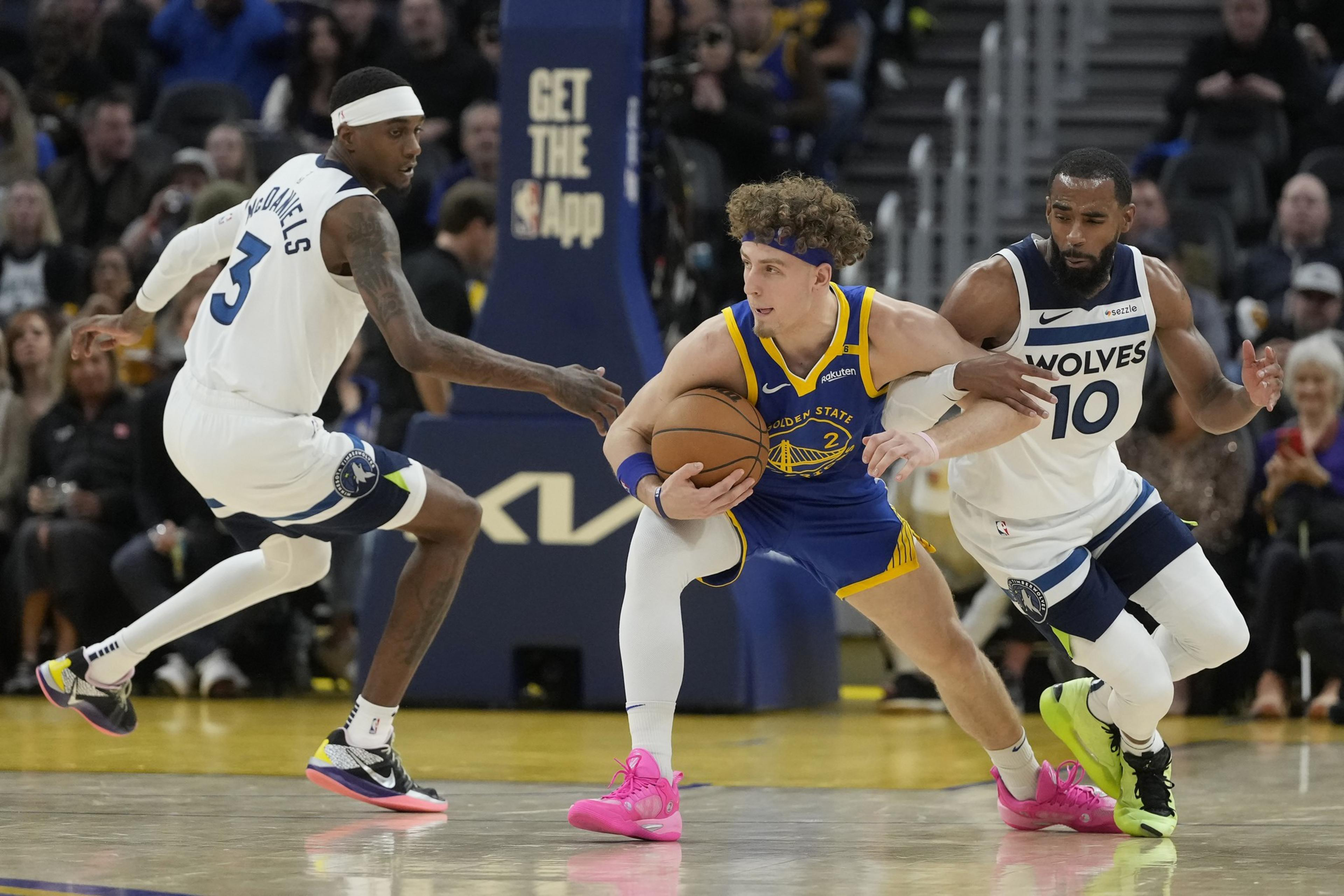 A basketball player in a blue and yellow uniform dribbles the ball while two players in white uniforms defend him on the court, with spectators in the background.