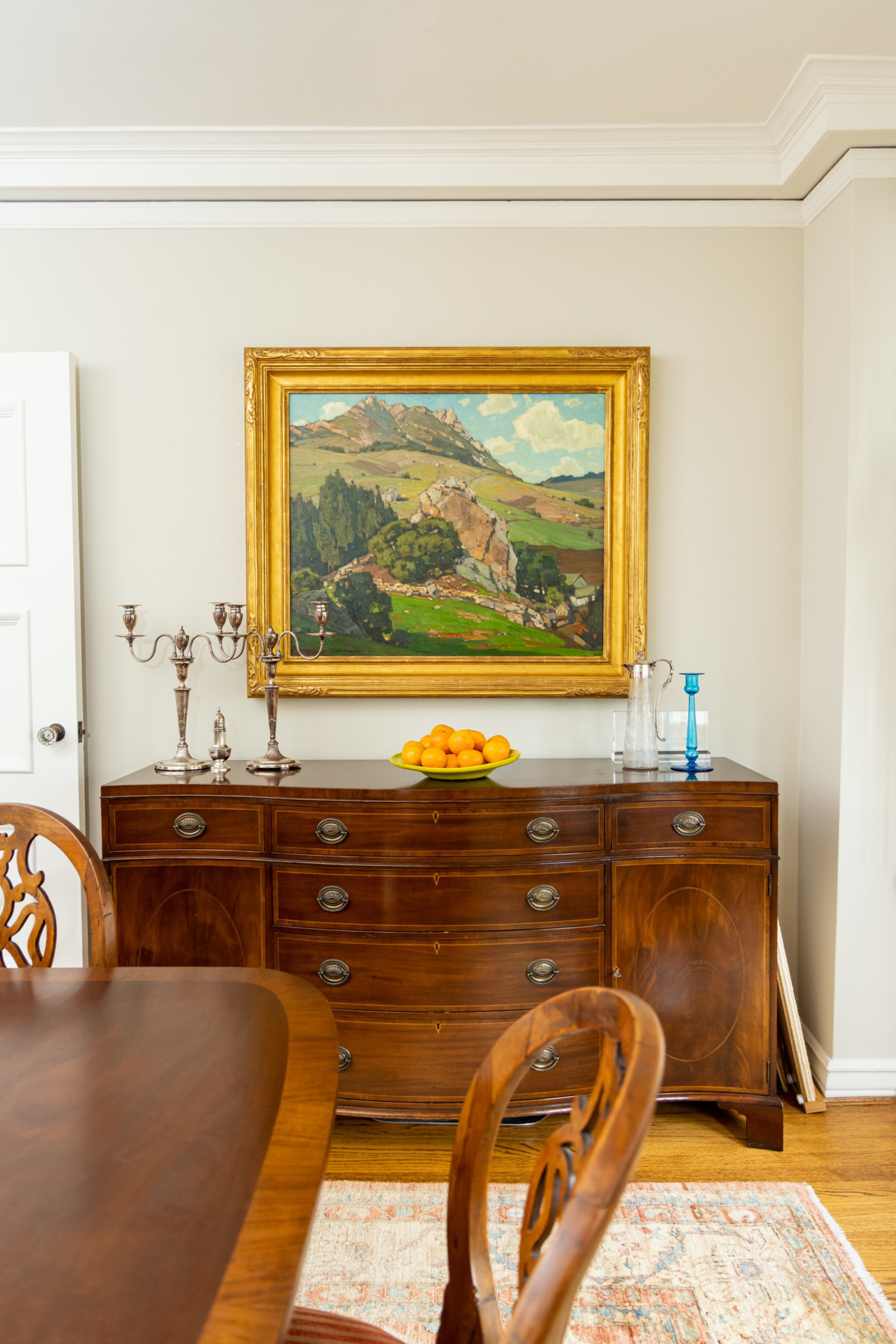 A wooden sideboard with silver candlesticks, a plate of oranges, glass bottles, and a blue candle holder sits beneath a framed landscape painting on a light wall.