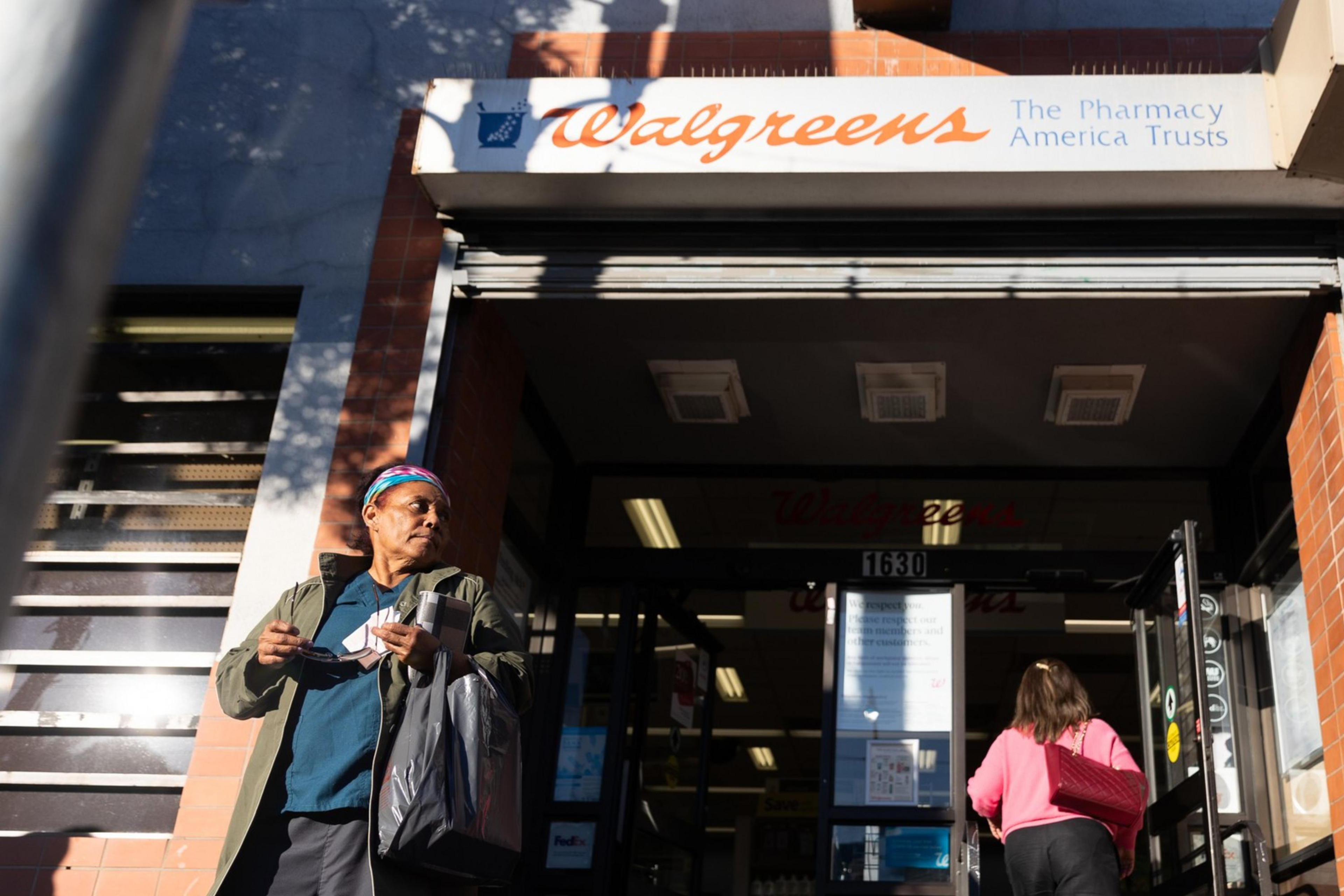 The image shows a Walgreens entrance with a person walking out holding a bag and another person entering the store. The building is brick with visible signage.