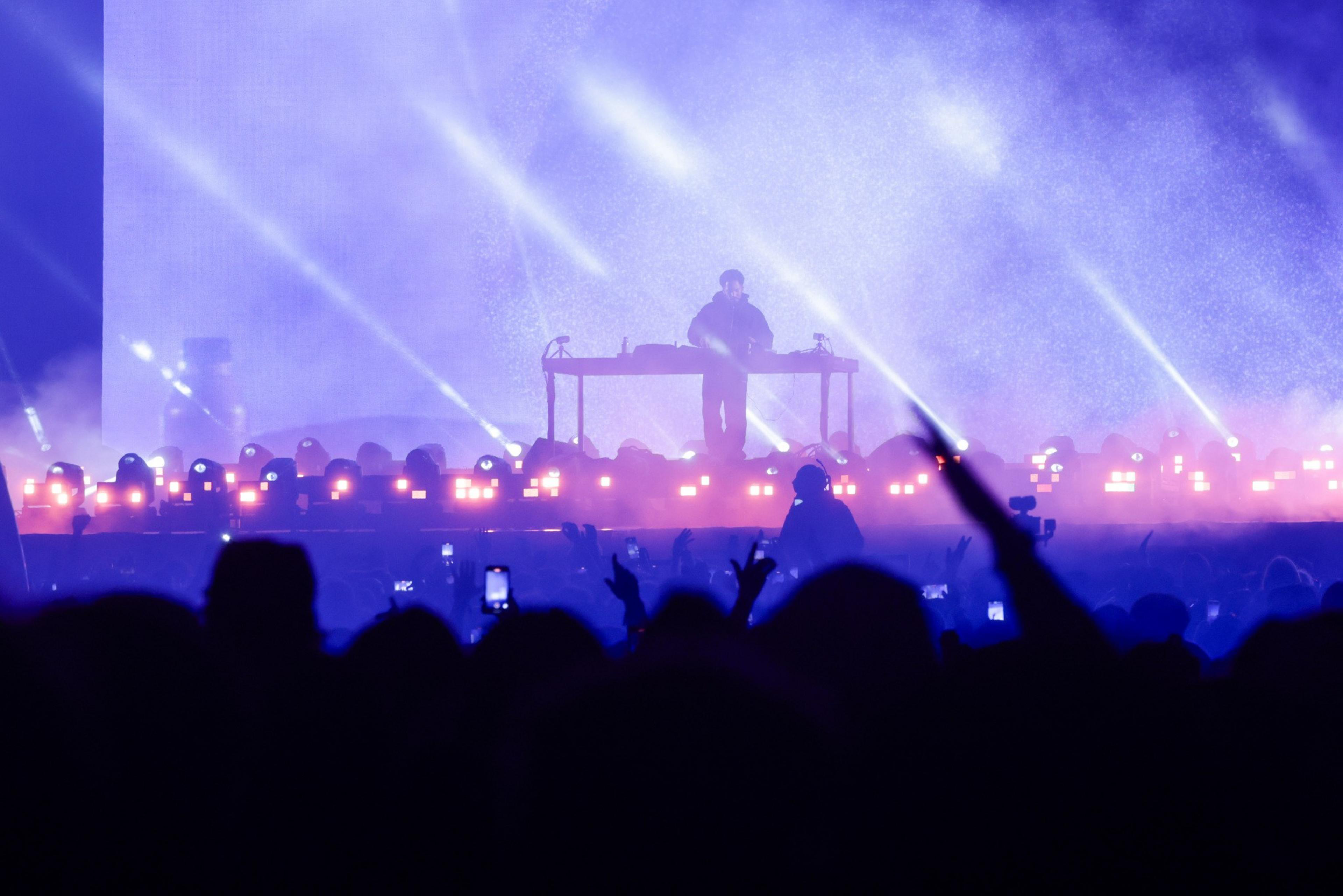 A DJ performs on a lit stage with bright beams and purple lighting as a crowd watches, some holding up phones and raising hands.