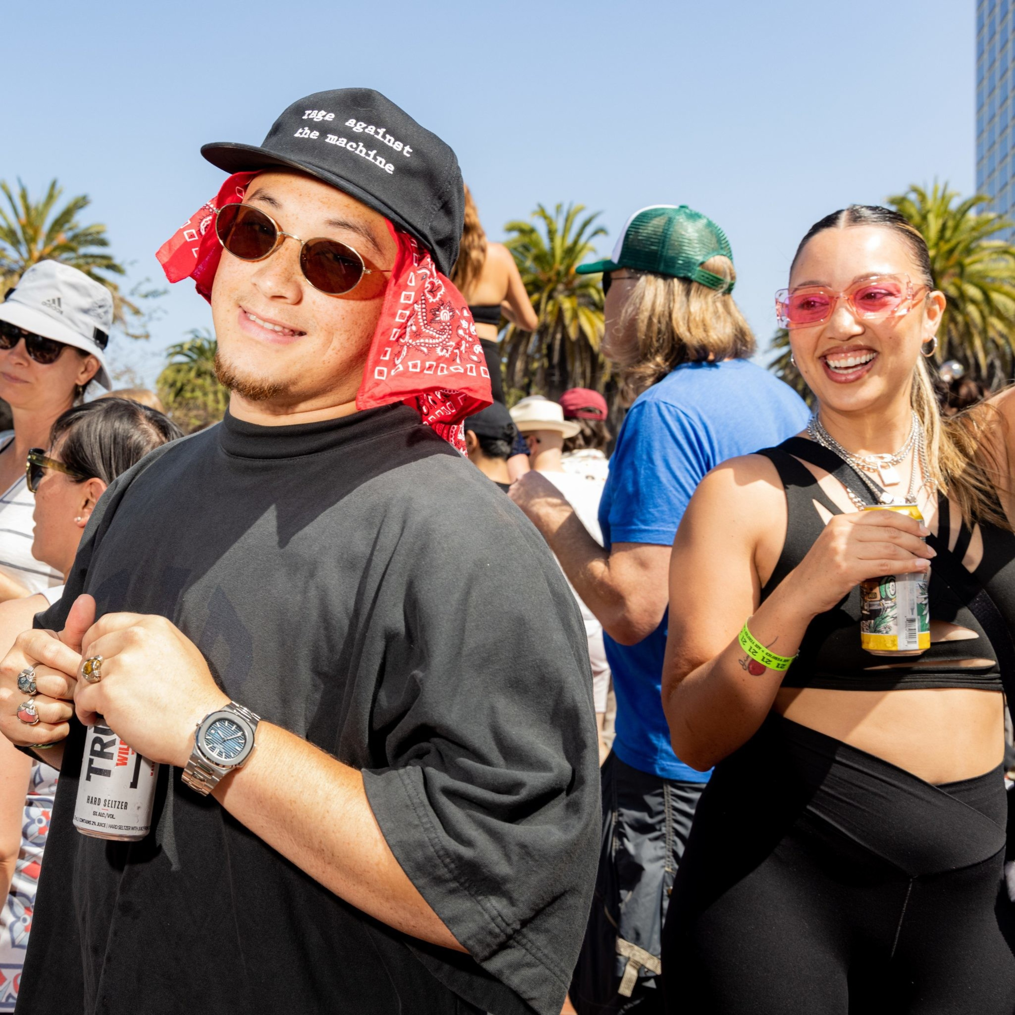 A smiling man in dark clothes and sunglasses holds a drink can, surrounded by an outdoor crowd under a clear sky. A woman nearby also smiles, holding a beverage.