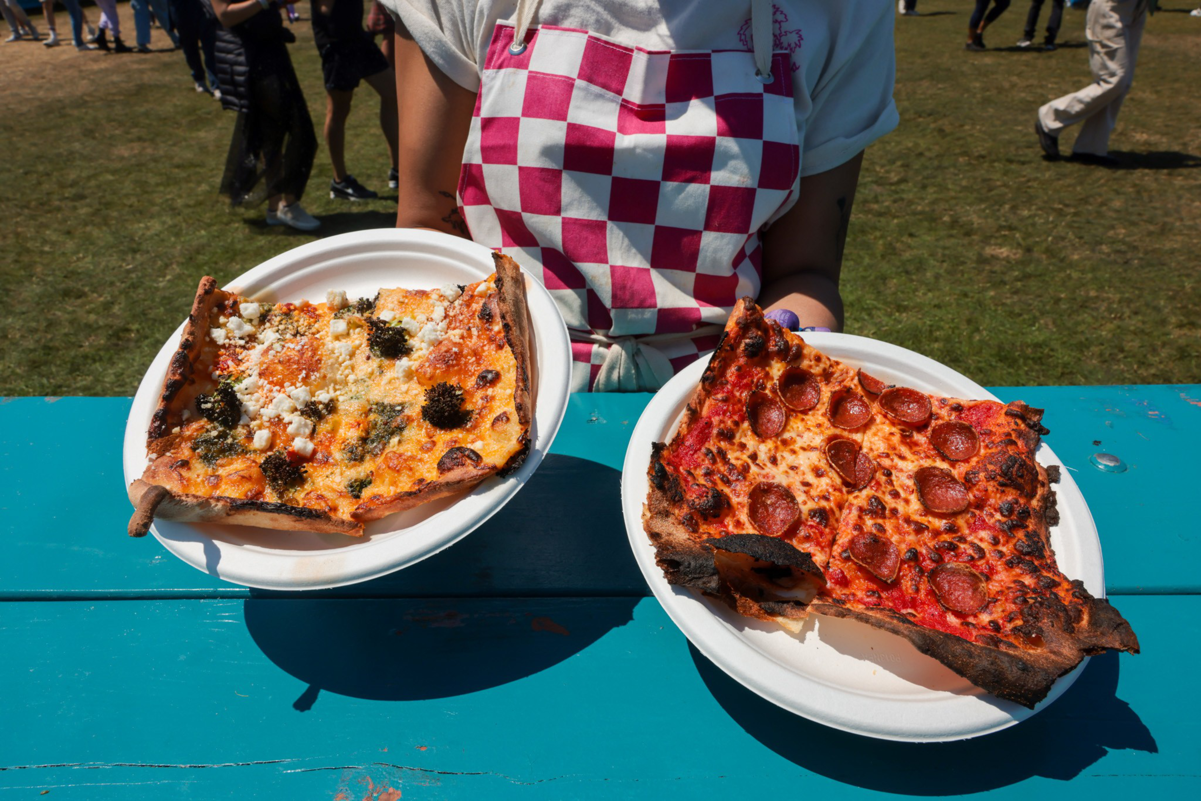 Two plates hold large, thin slices of pizza—one with pepperoni, the other with cheese and herbs—held by a person wearing a pink-and-white checkered apron.