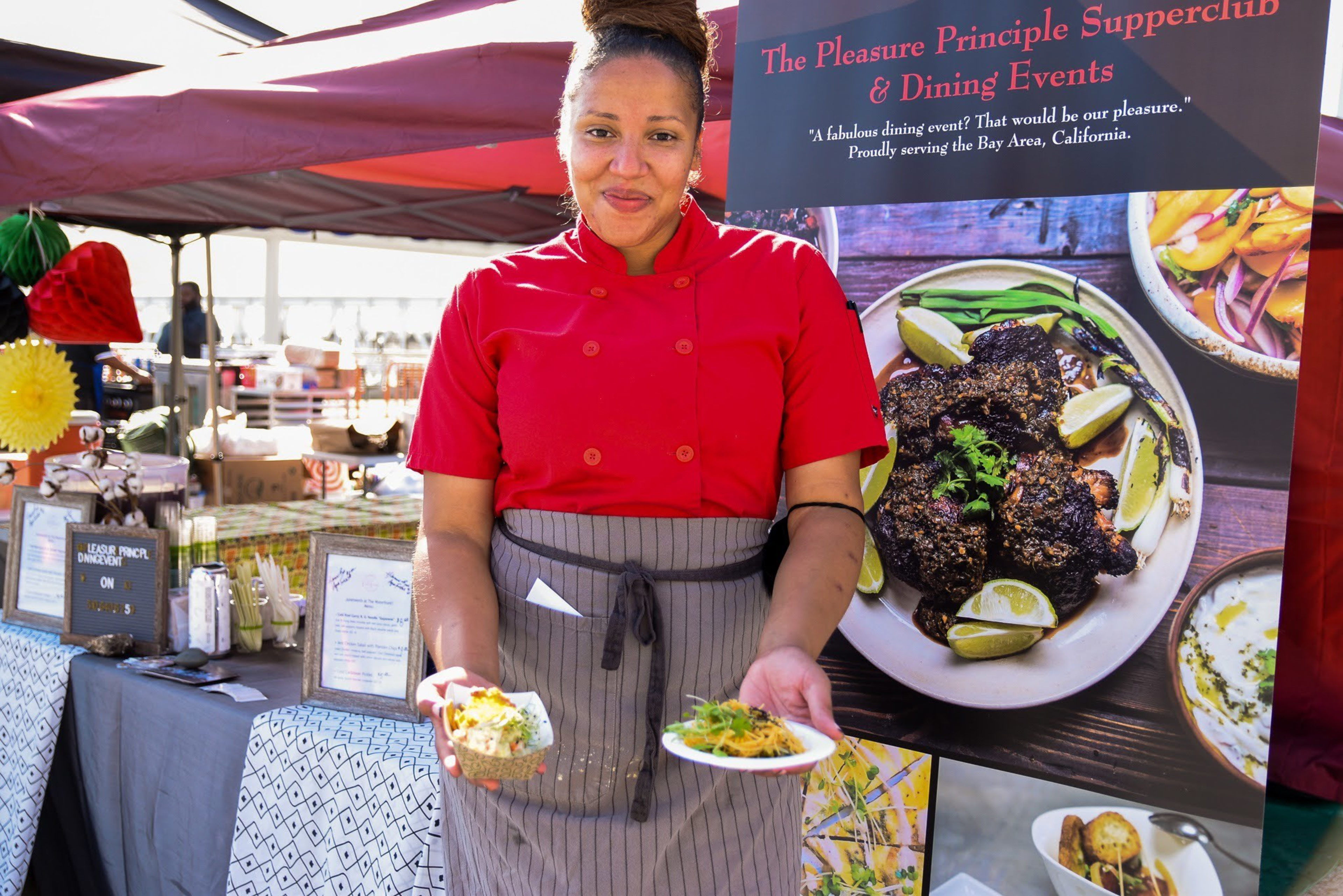A woman in a red chef coat and striped apron holds two plated dishes at an outdoor market stall with a sign for The Pleasure Principle Supperclub.