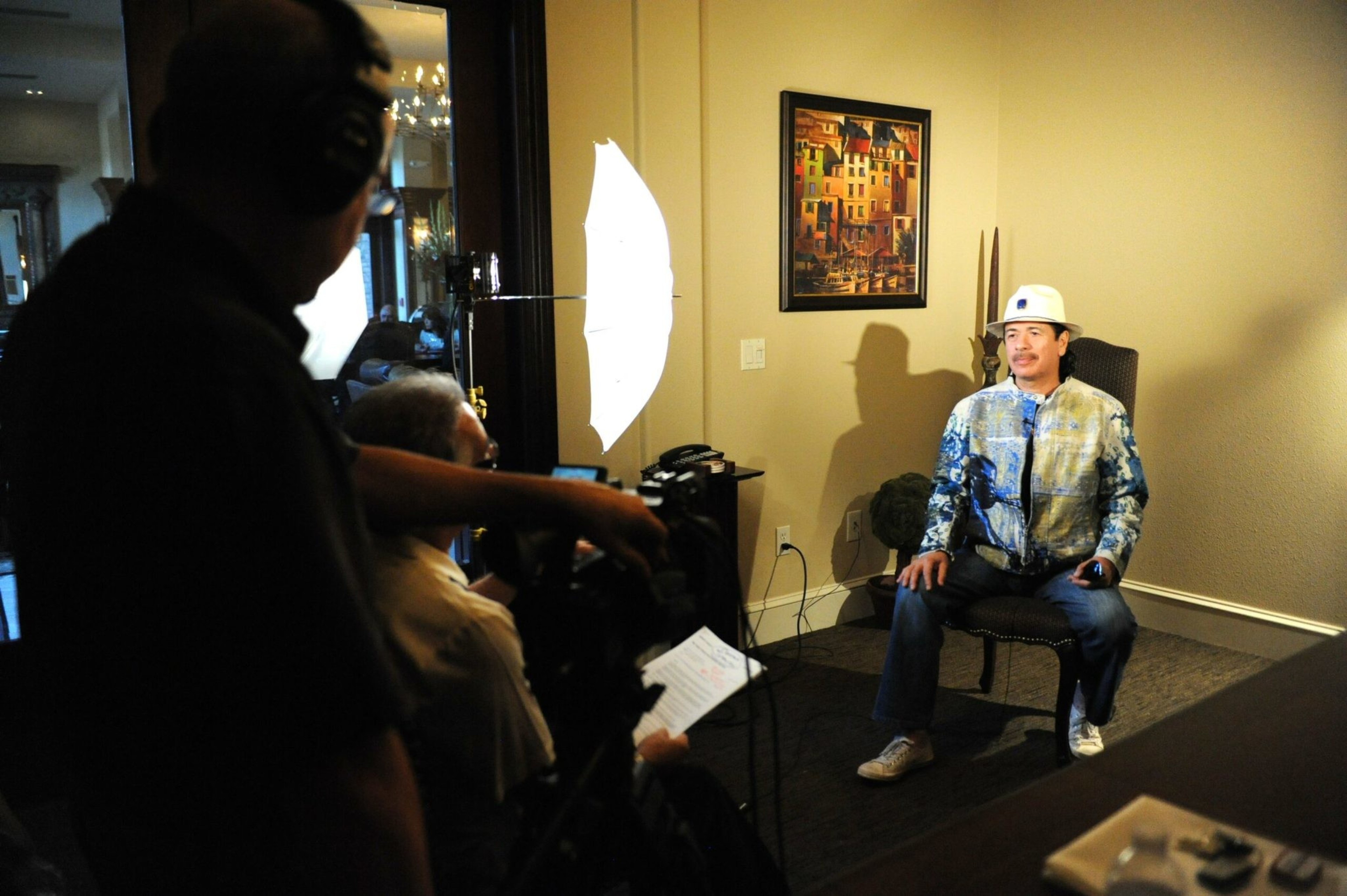 A man wearing a white hard hat and colorful jacket sits on a chair, illuminated by studio lights while a crew films him indoors.