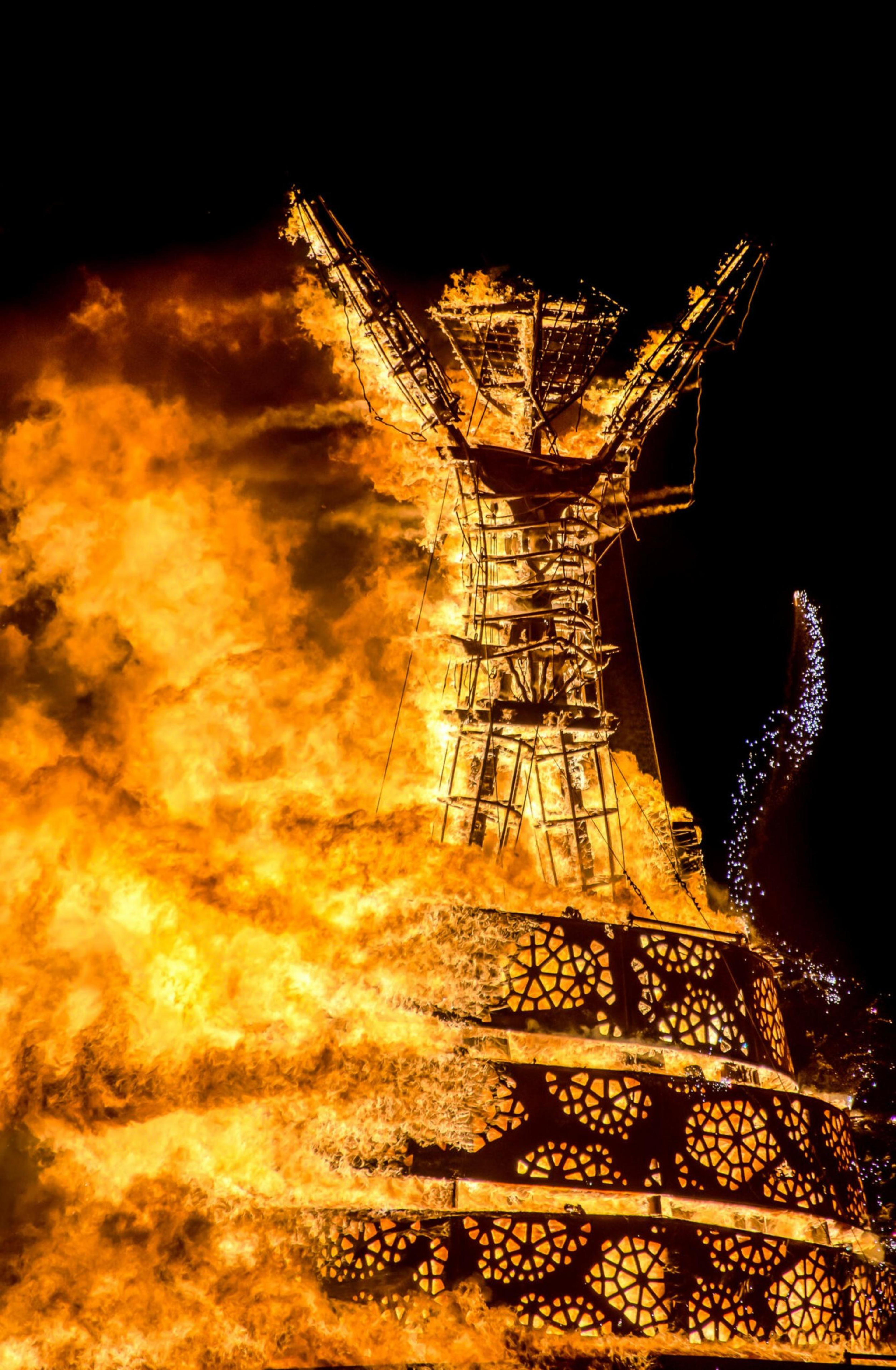 A large wooden structure resembling a human figure is engulfed in bright orange flames against a dark sky, creating a dramatic, fiery spectacle.