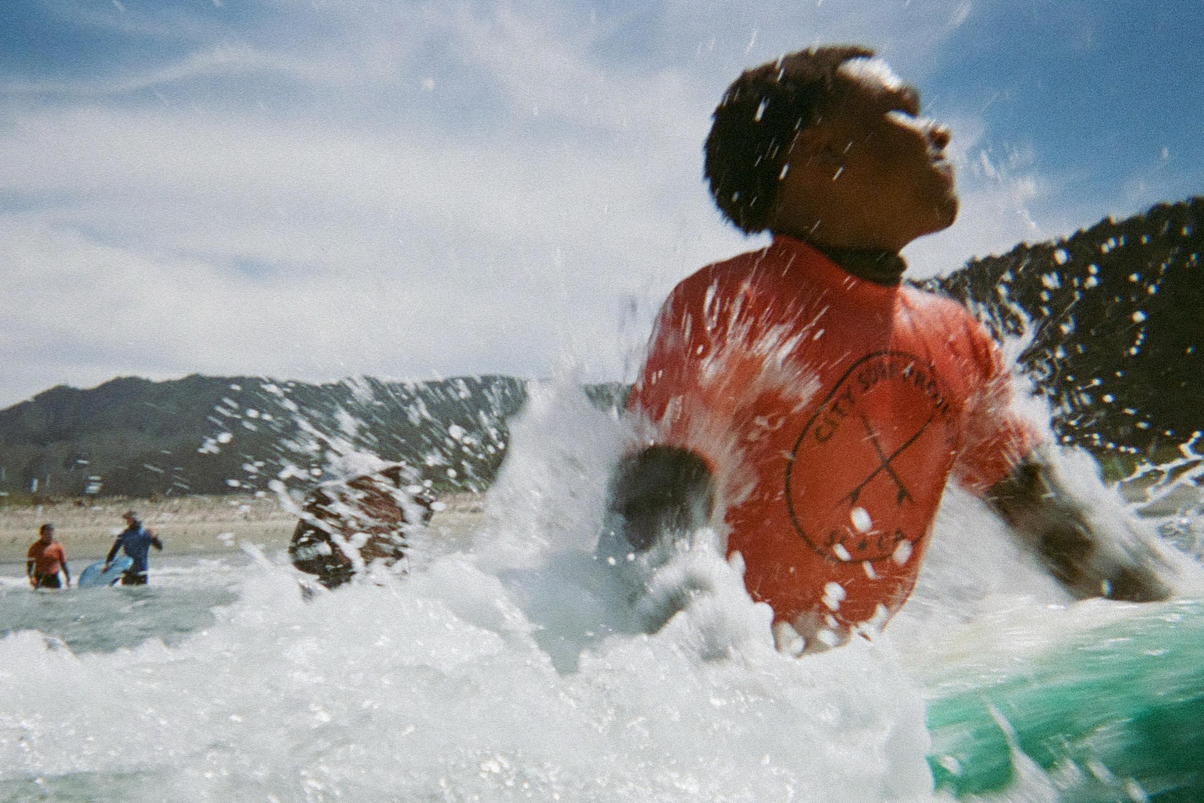 A person in a red shirt is in the foreground, partially submerged in a splash of ocean waves. In the background, others stand in the water near the shore.
