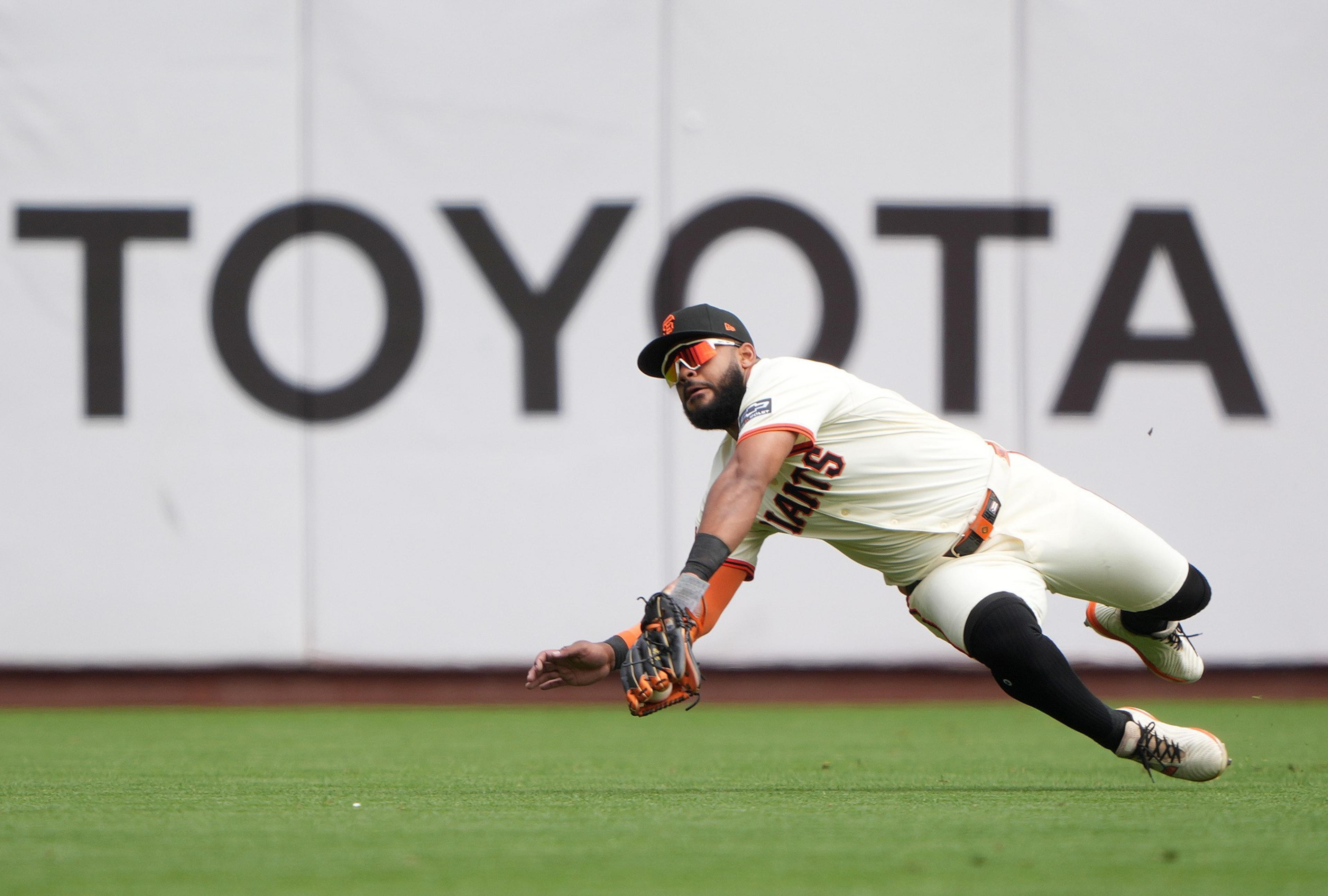 A baseball player in a San Francisco Giants uniform dives to catch a ball near an outfield wall with a large "TOYOTA" sign.
