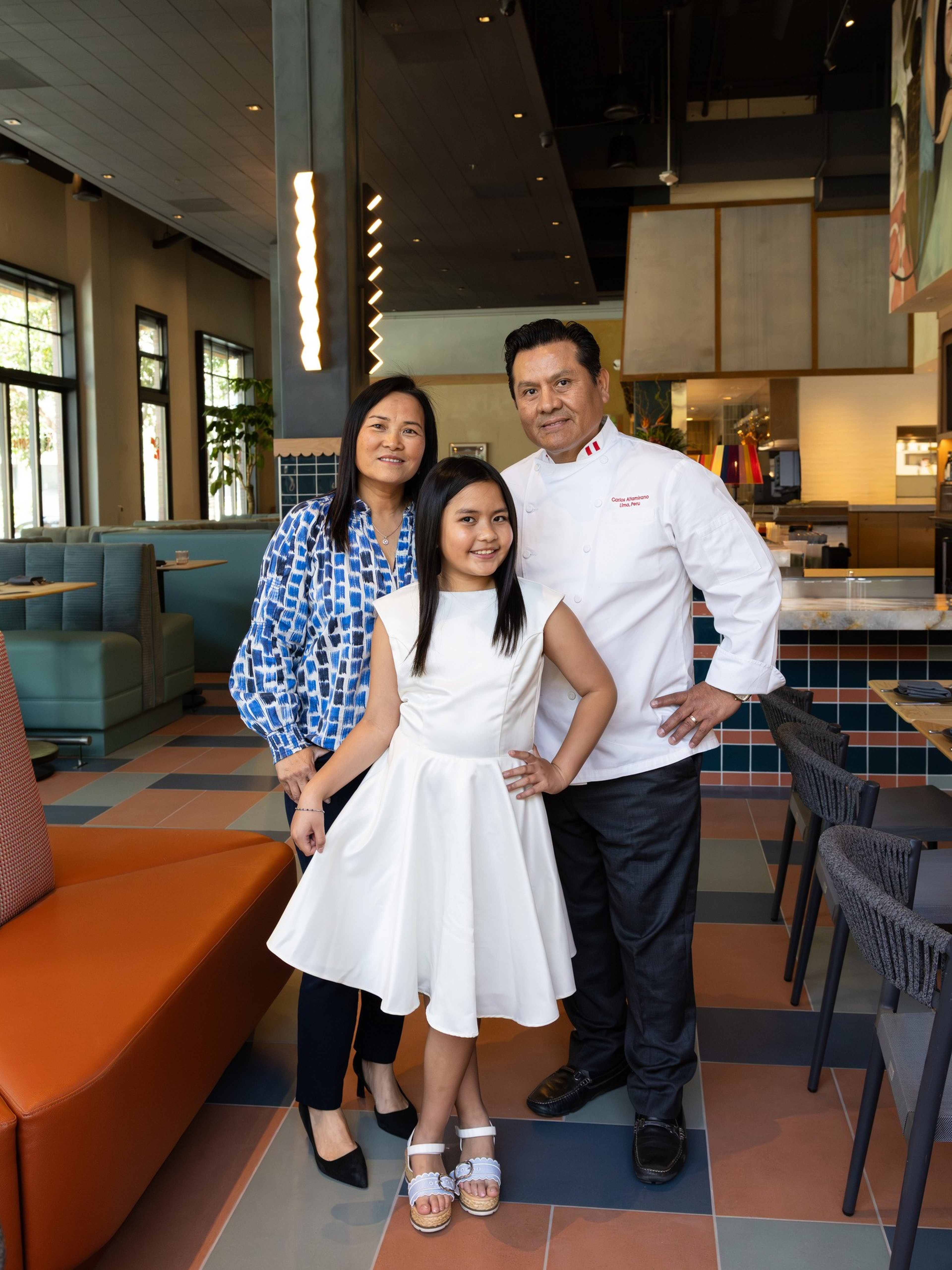 A man in a chef’s uniform, a woman in a blue patterned blouse, and a girl in a white dress pose together inside a modern restaurant.