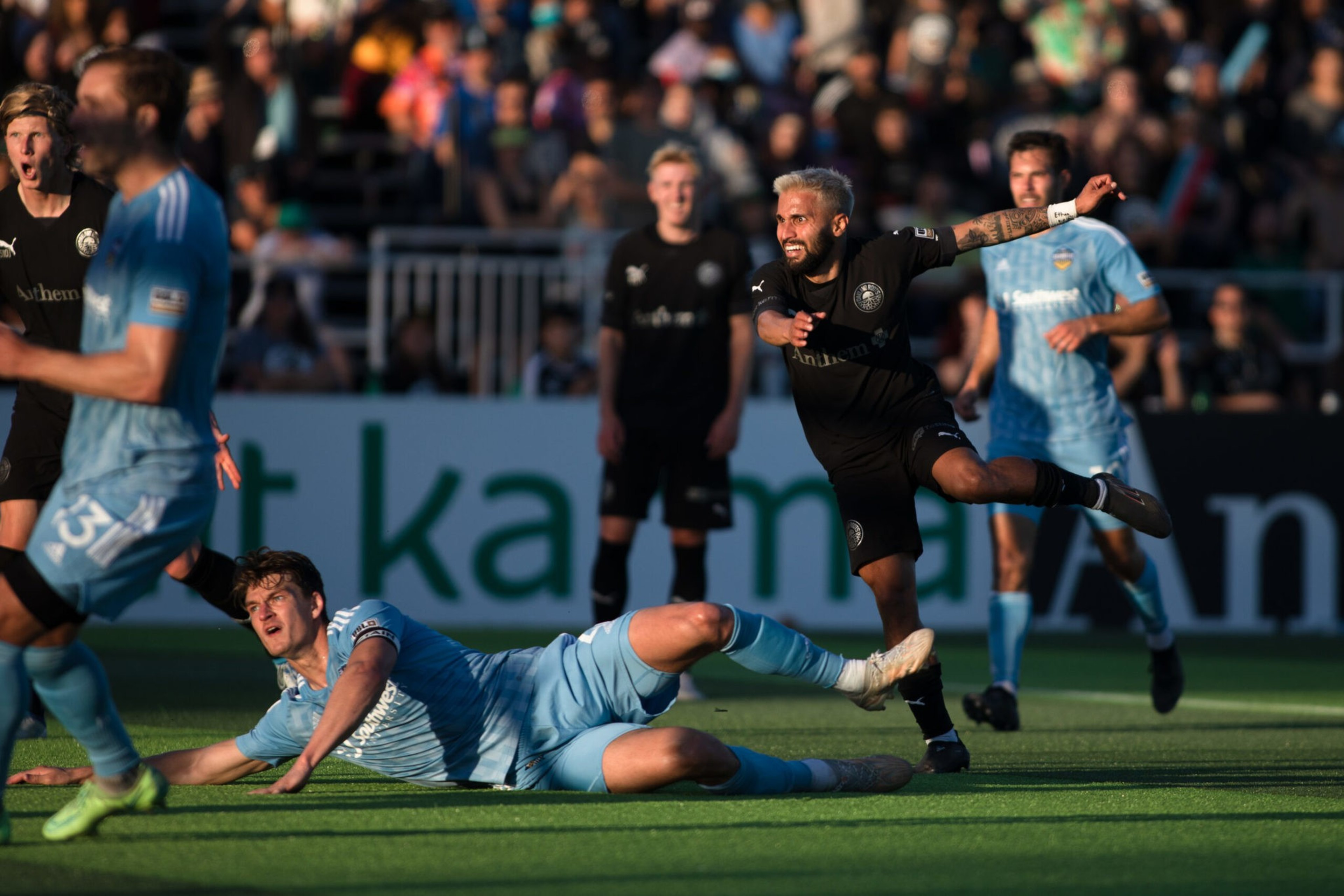 A soccer player in a black jersey kicks the ball while a player in a light blue jersey slides on the ground attempting to block it.