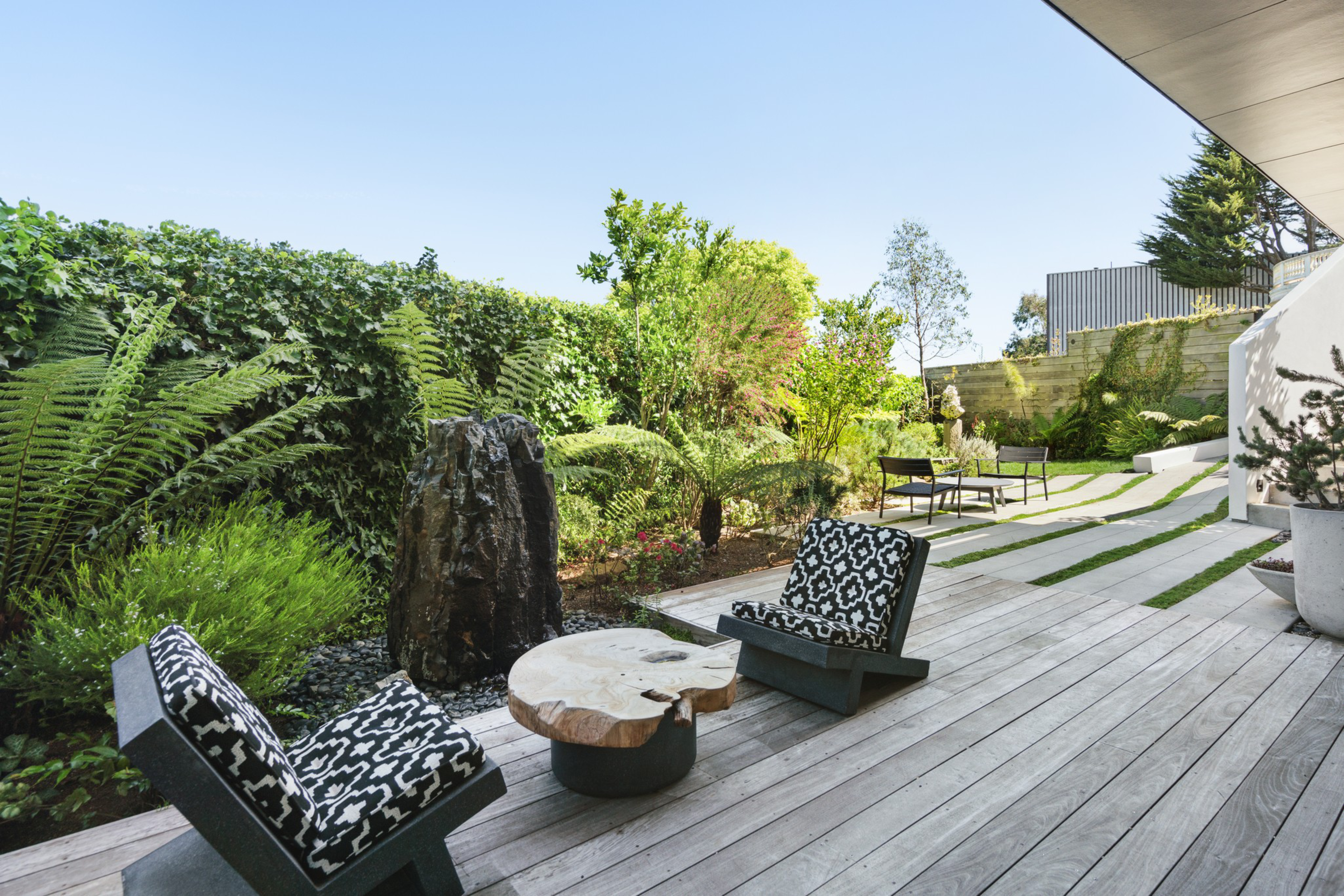 A wooden deck features two modern black chairs with patterned cushions and a round natural wood coffee table, surrounded by lush green plants and a small water fountain.