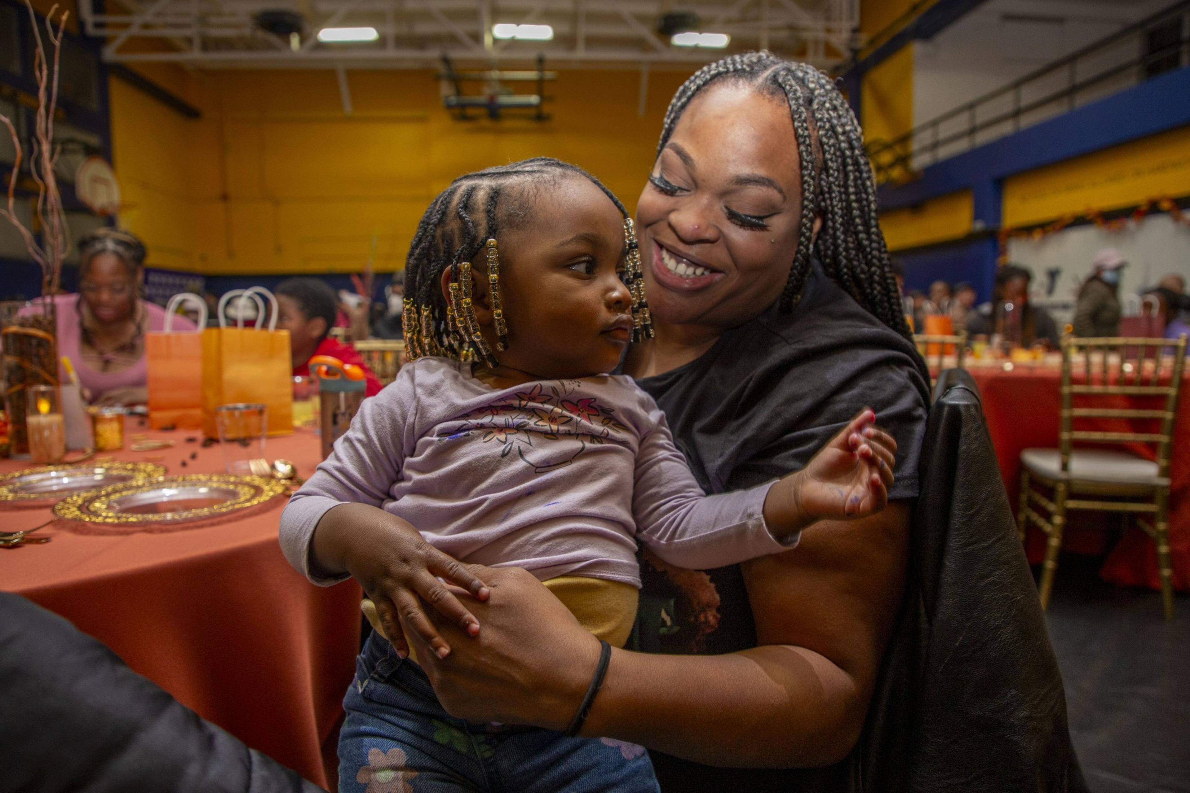 A smiling woman holds a young child with braided hair in a lively indoor setting with decorated tables and orange-themed gifts in the background.