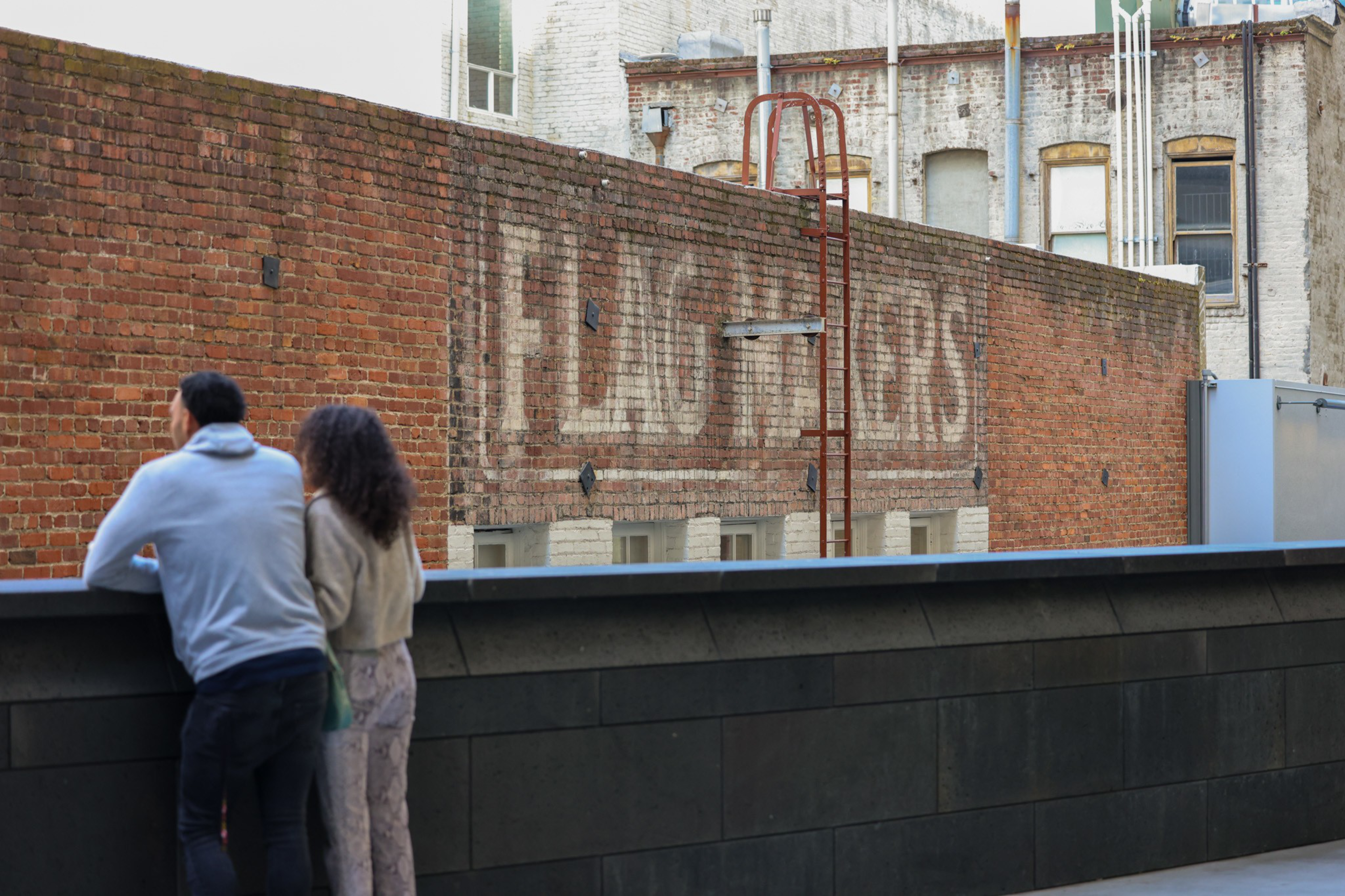 Two people lean against a railing looking at a brick wall with a faded painted advertisement that reads "RED CROSS FLOORS."