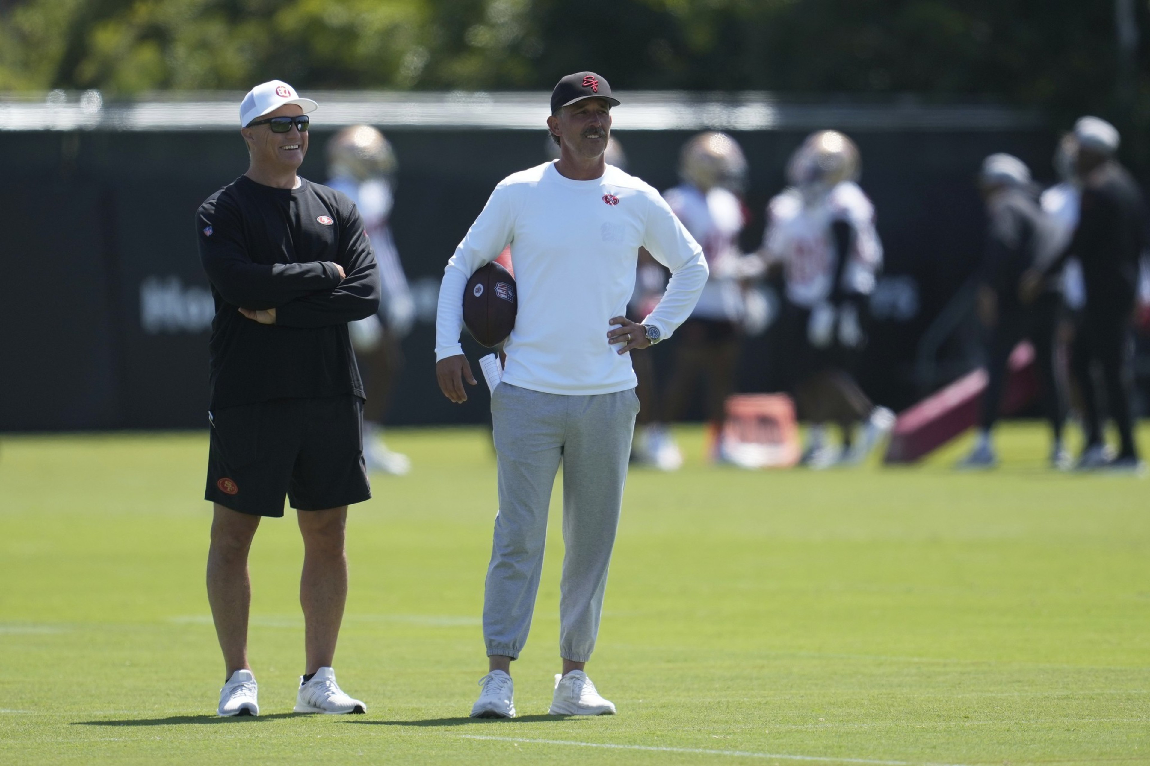 Two men stand on a grassy field during a practice. One wears a black outfit and cap, the other a white top and gray pants, holding a football. Others practice in the background.