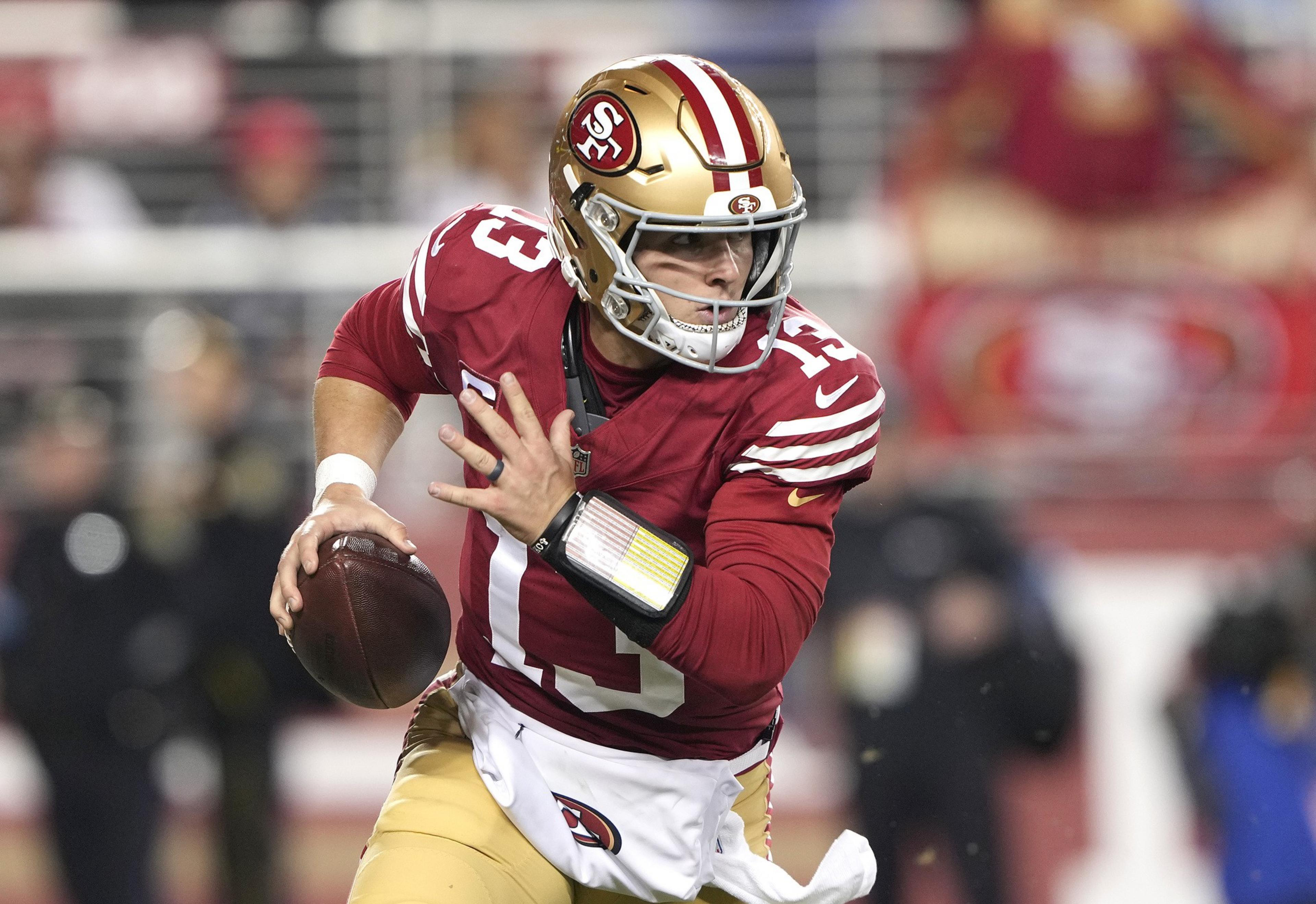 A football player in a red jersey and gold pants runs with the ball, focused and poised. He's wearing a helmet with the number 13 and a team's logo.
