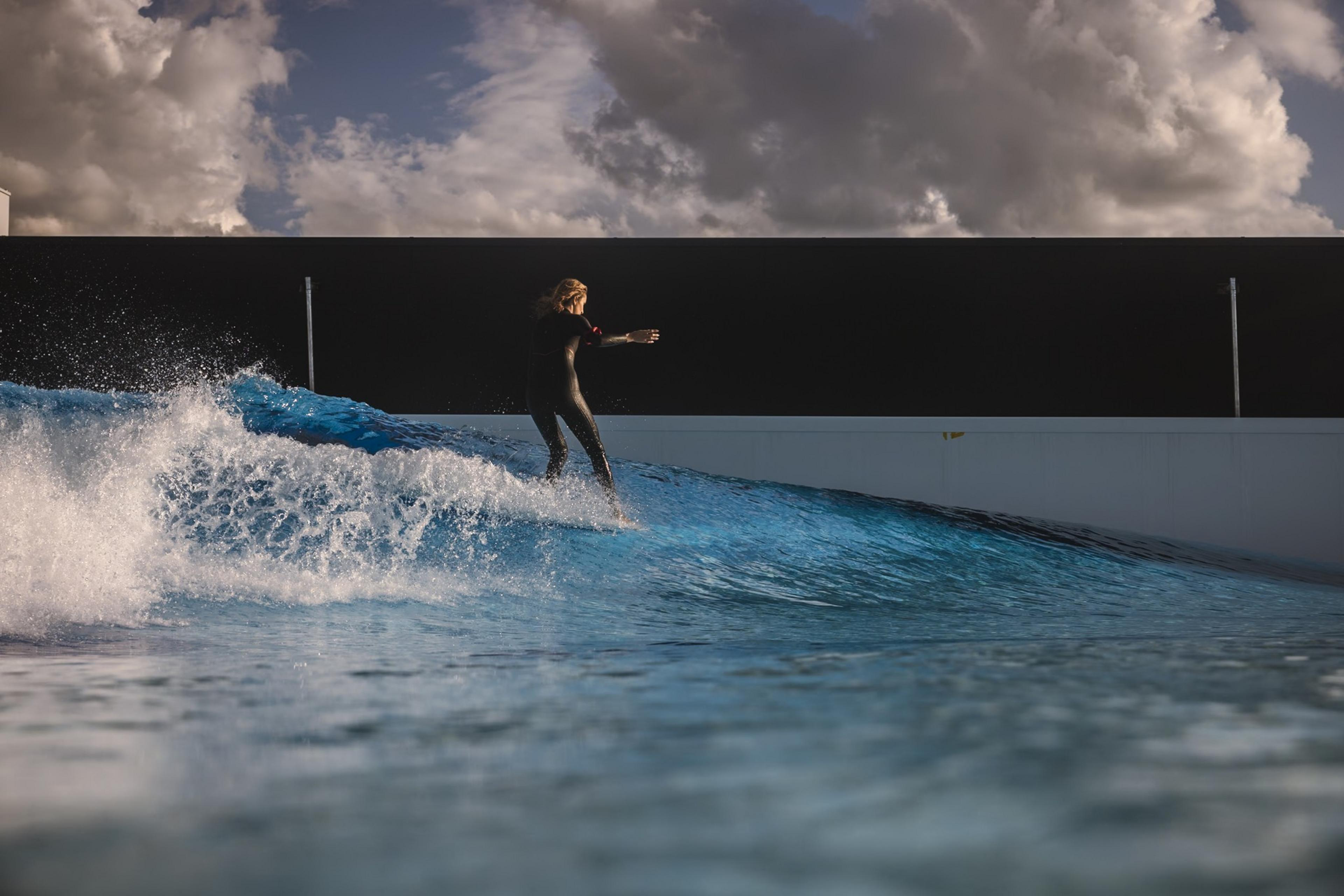 A person in a wetsuit surfs on a bright blue artificial wave under a dramatic cloudy sky, against a modern structure's backdrop.