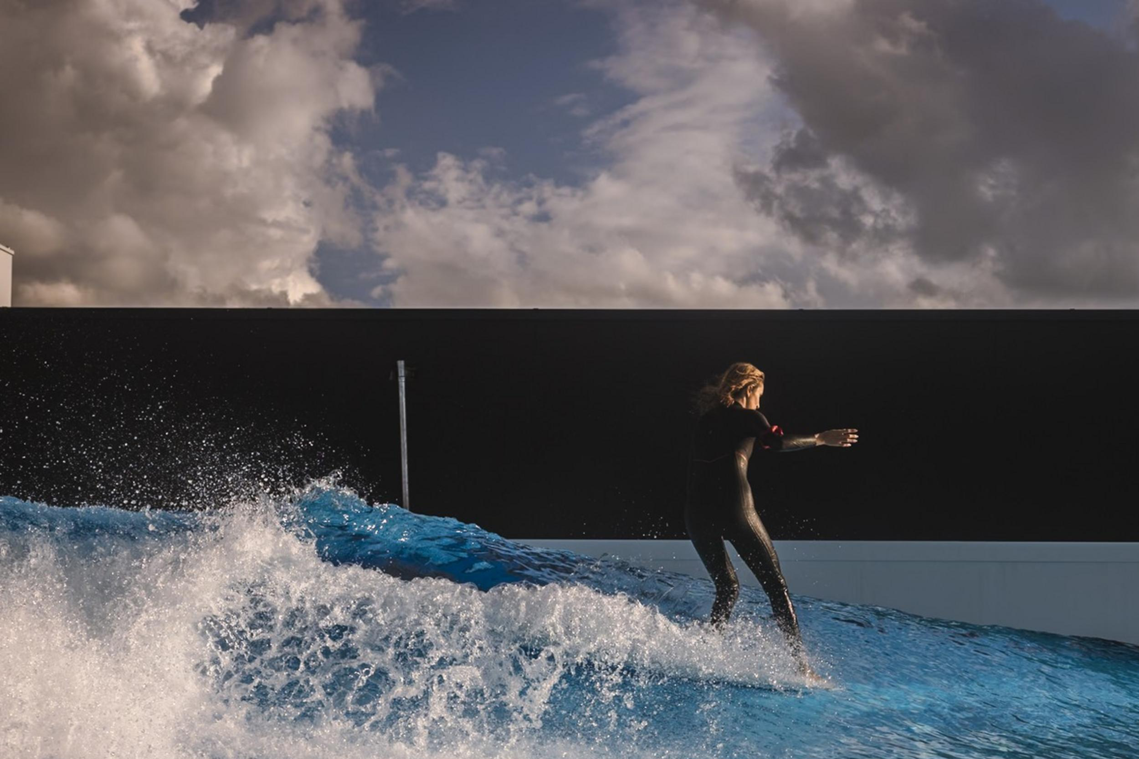 A person in a wetsuit surfs on a bright blue artificial wave under a dramatic cloudy sky, against a modern structure's backdrop.