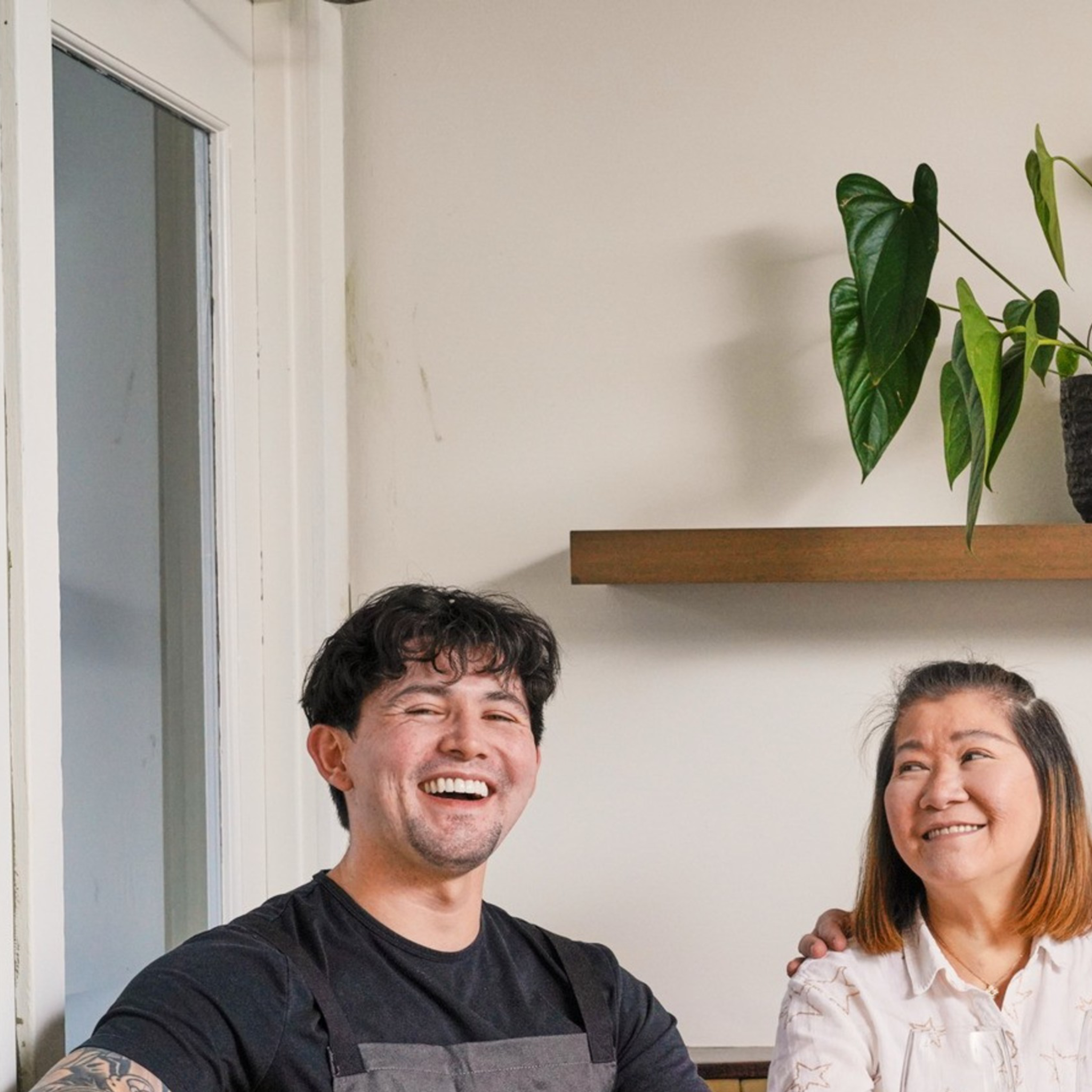 Three people sit at a wooden table with wine glasses, smiling. A plant is on a shelf behind them, and there are flowers on the table. The setting is cozy and bright.
