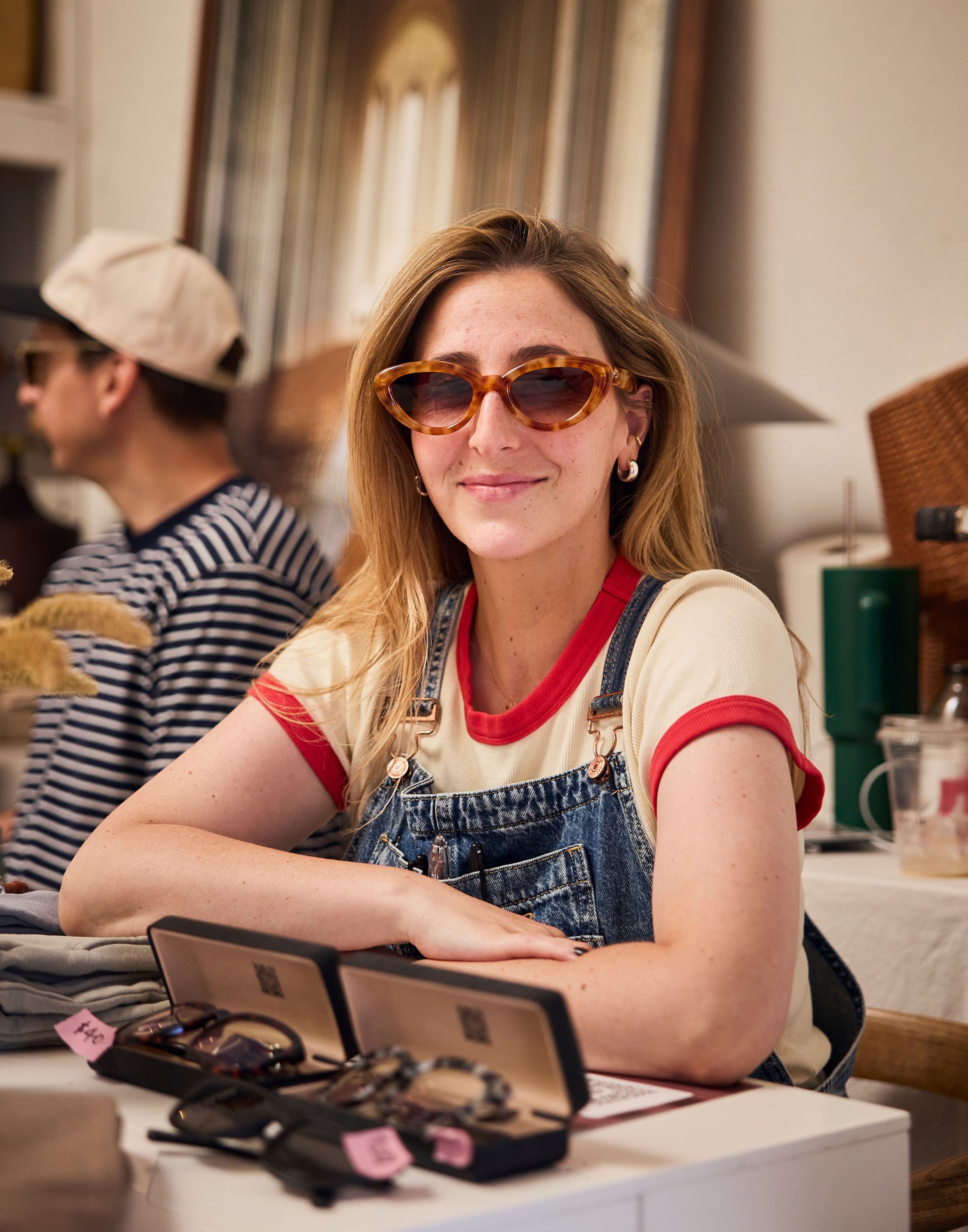 A woman wearing tortoiseshell sunglasses, a red and white t-shirt, and denim overalls sits at a table with multiple pairs of eyeglasses displayed in front of her.