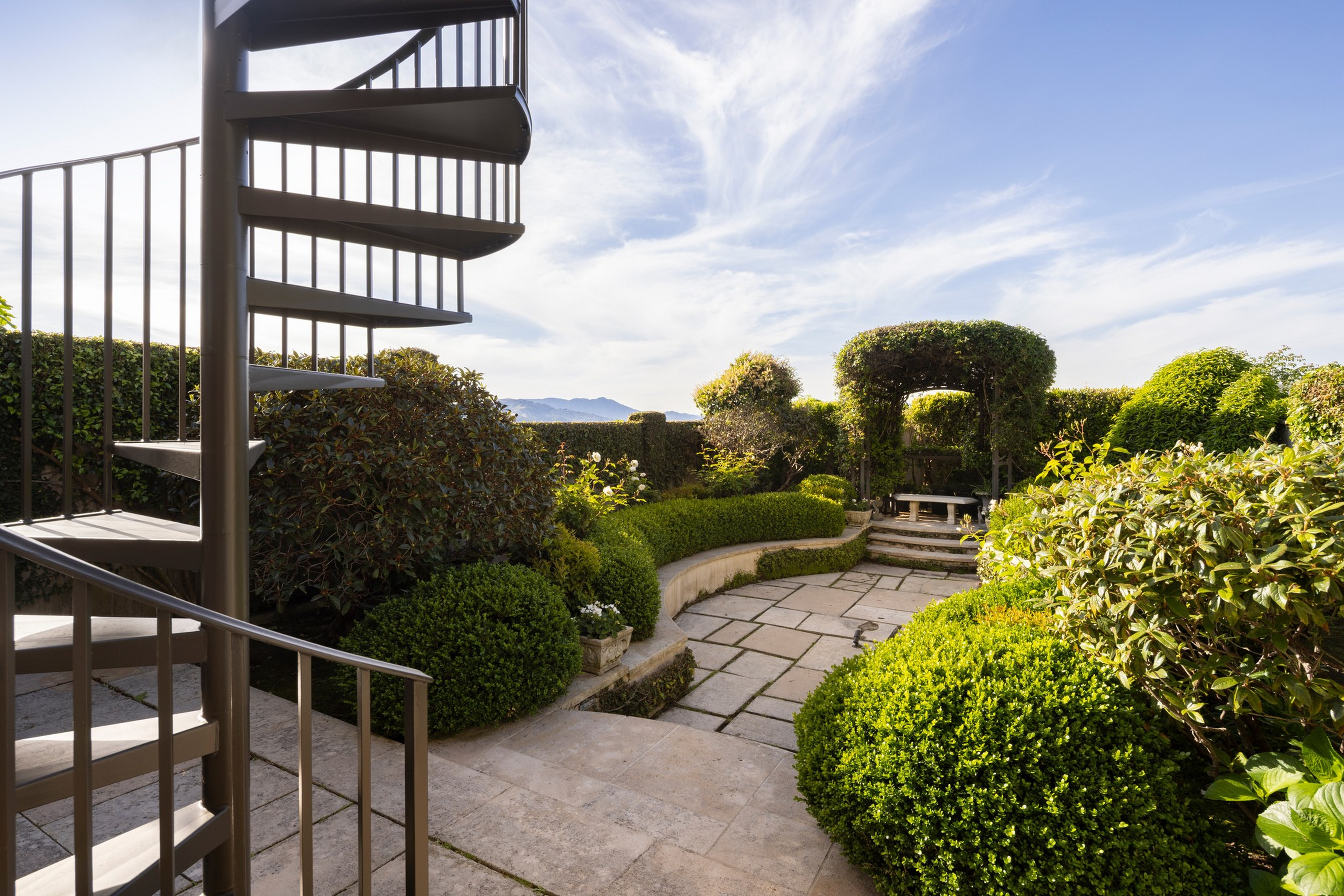 A spiral metal staircase overlooks a stone patio with manicured bushes, trimmed hedges, and a leafy archway leading to a shaded seating area.