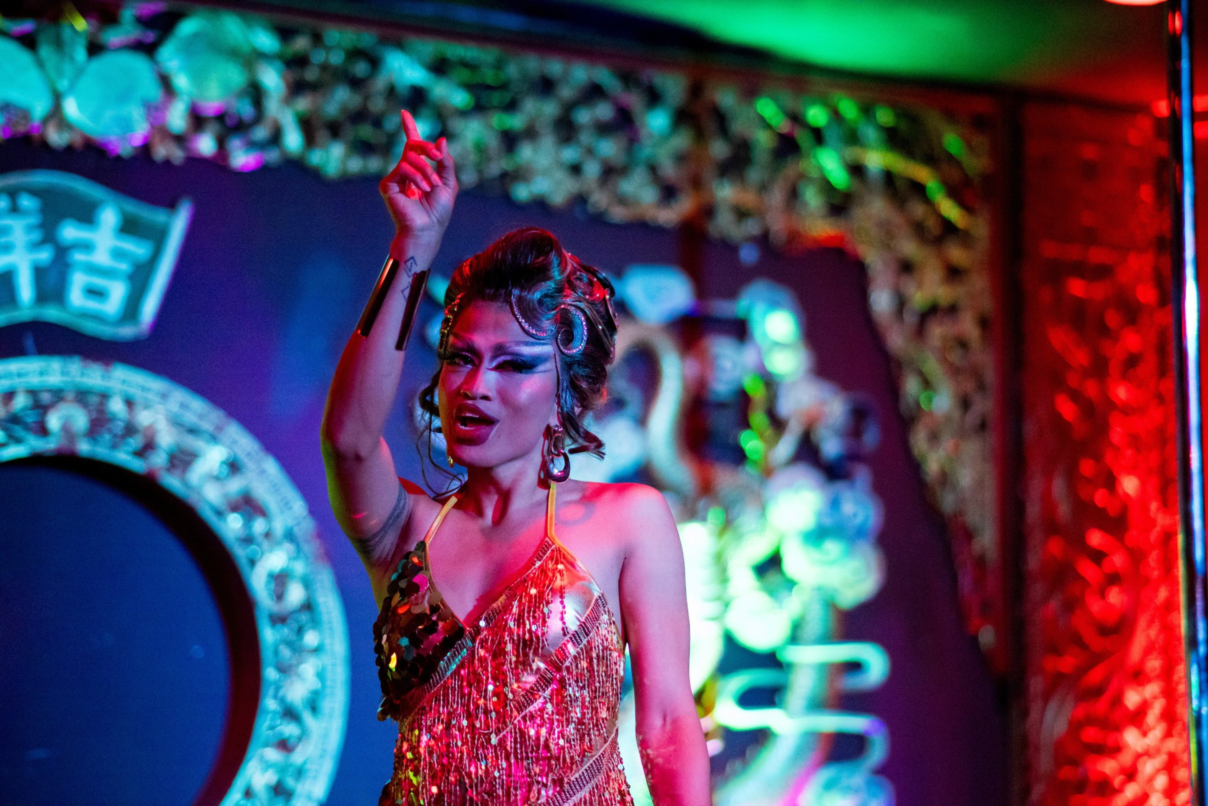 A performer in a glittery red dress gestures on stage, surrounded by vibrant lights and intricate decorations in a lively, colorful setting.