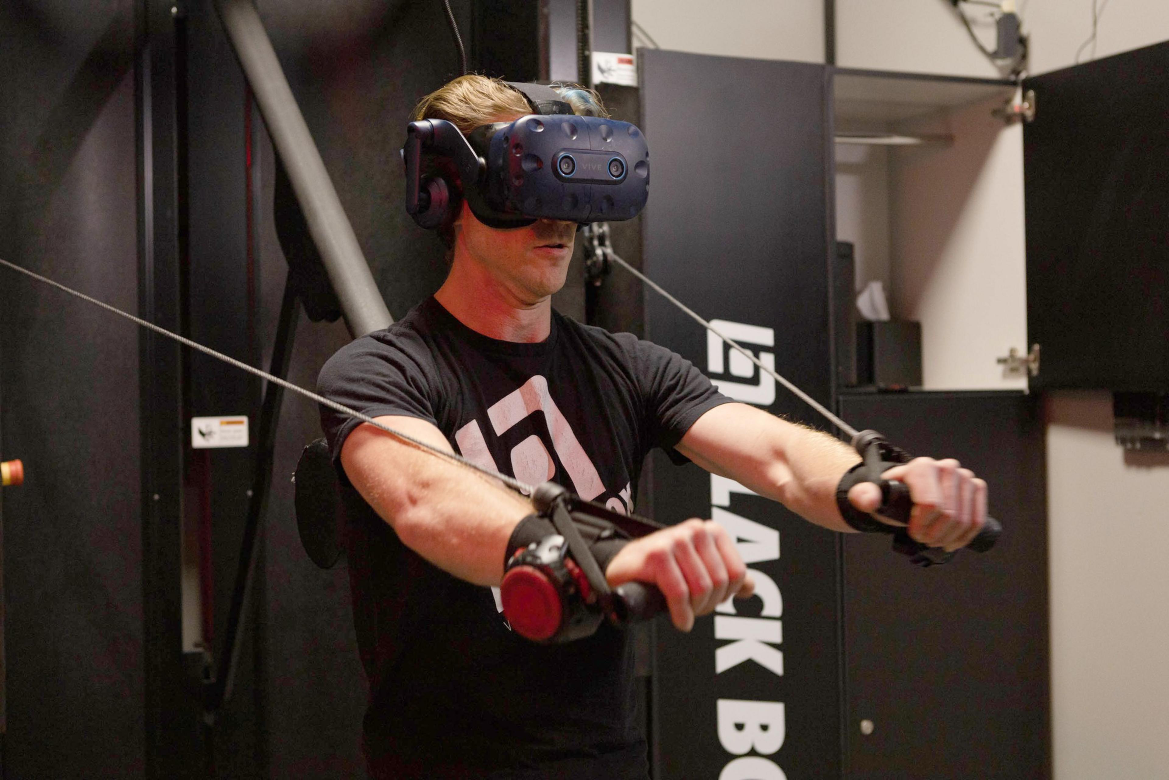 A man wearing a virtual reality headset uses hand controllers attached to cables, appearing to interact with a VR fitness or training setup.