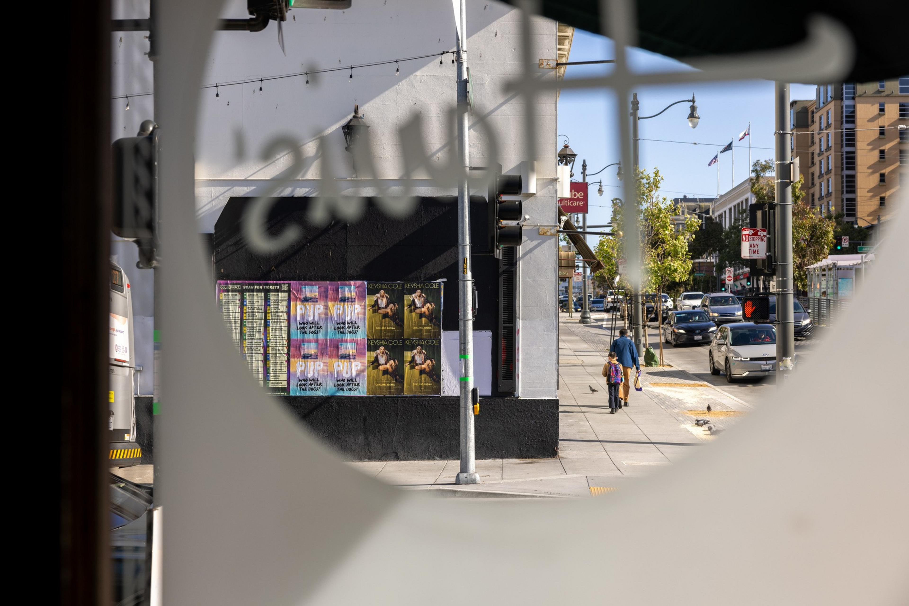 A city street view through a circular window, showing people walking, cars, and posters on a building. Traffic lights and trees are visible under a clear sky.