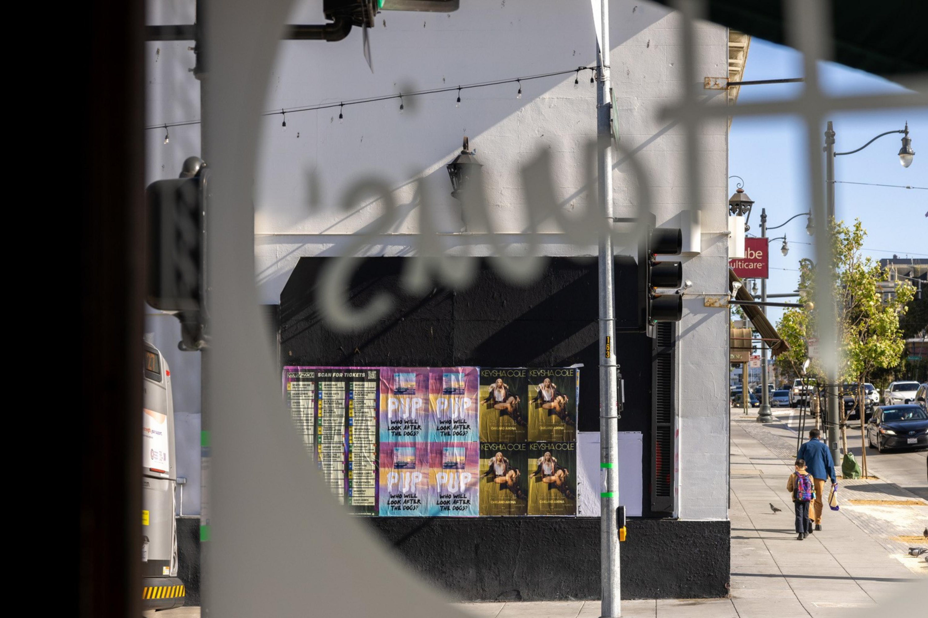 A city street view through a circular window, showing people walking, cars, and posters on a building. Traffic lights and trees are visible under a clear sky.