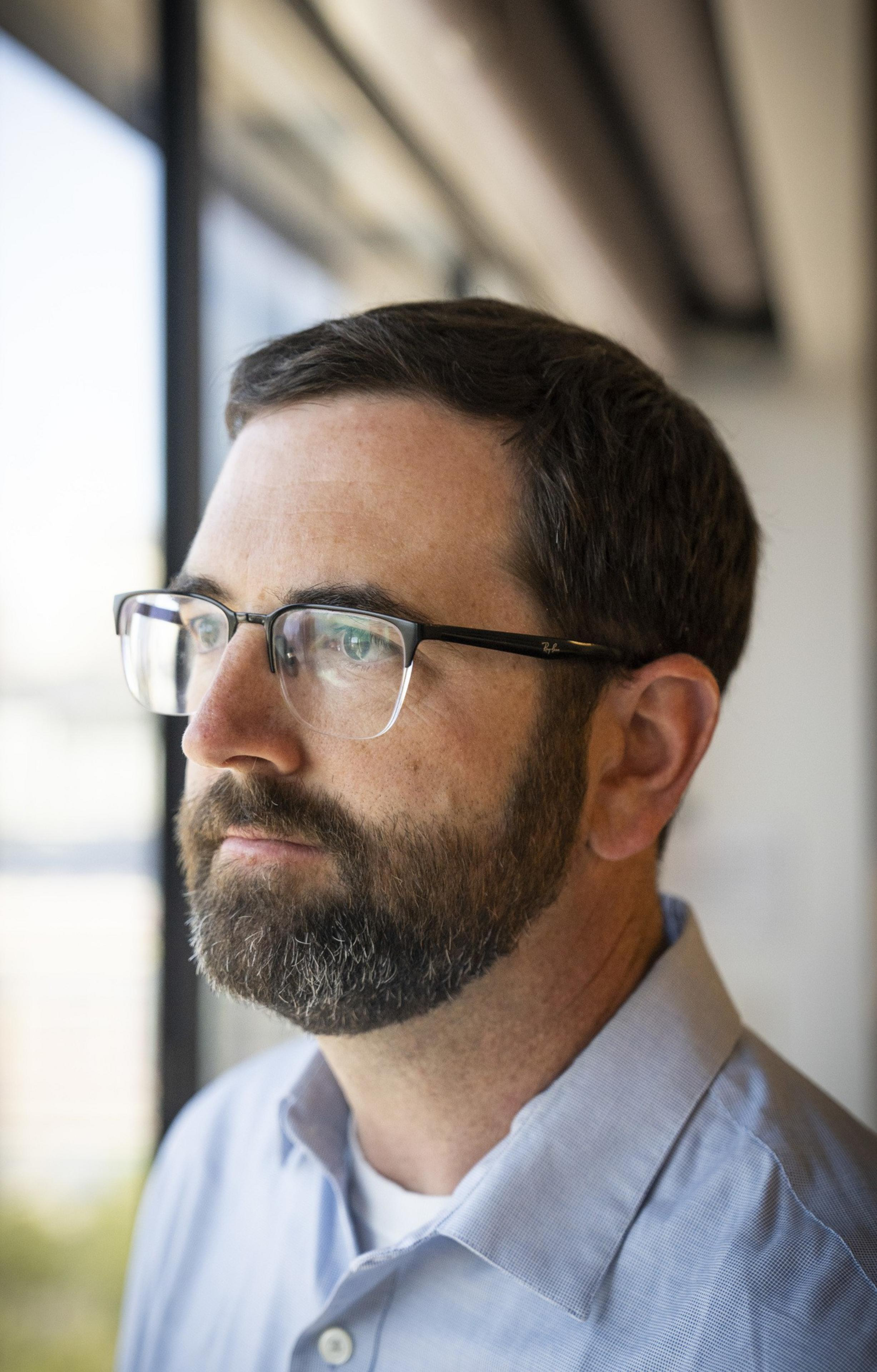 A bearded man wearing glasses and a light blue shirt looks pensively out a window, with soft natural light illuminating his profile.