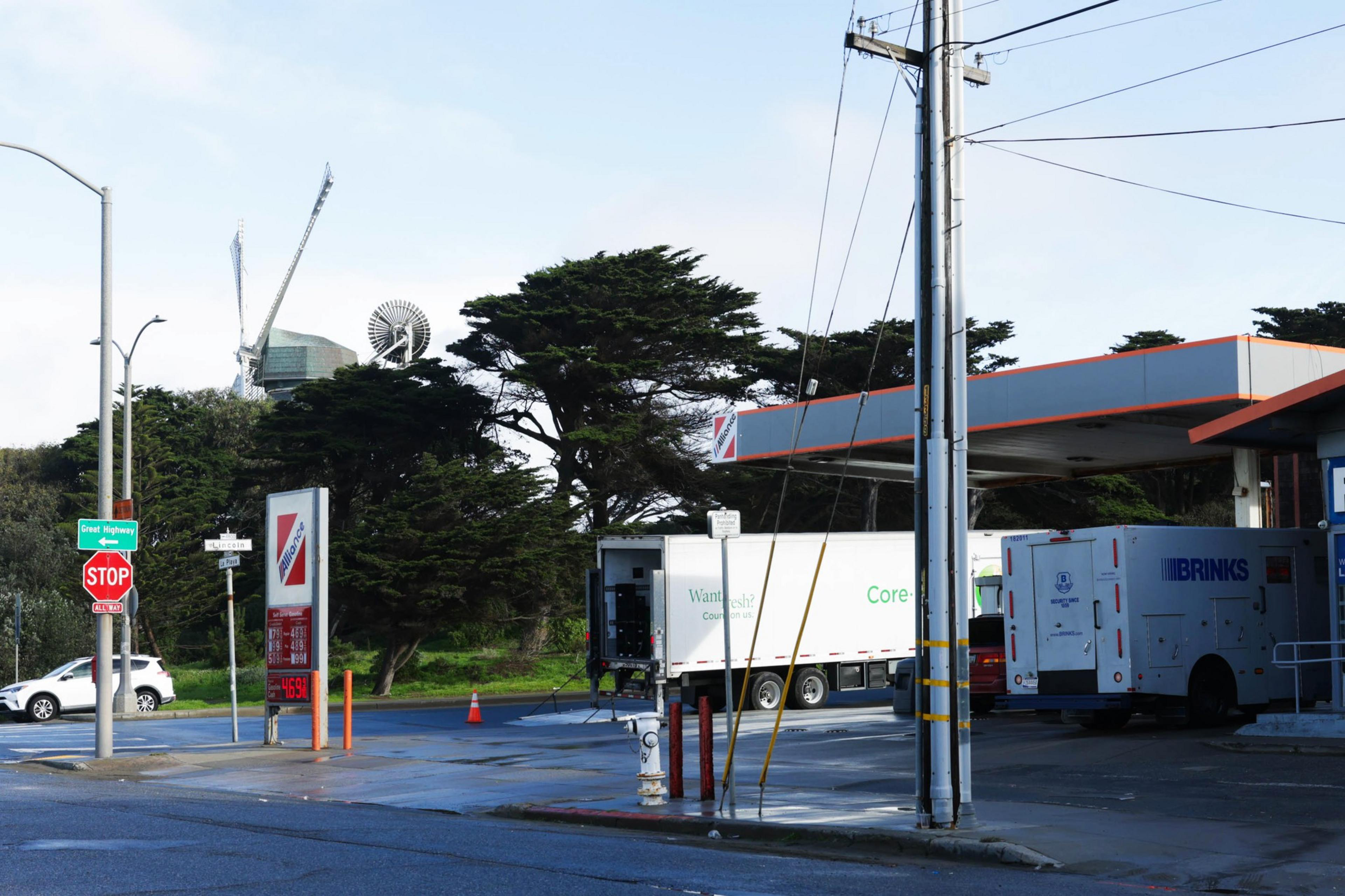 A street scene with a gas station, a Brinks security truck, and construction cranes against a blue sky.