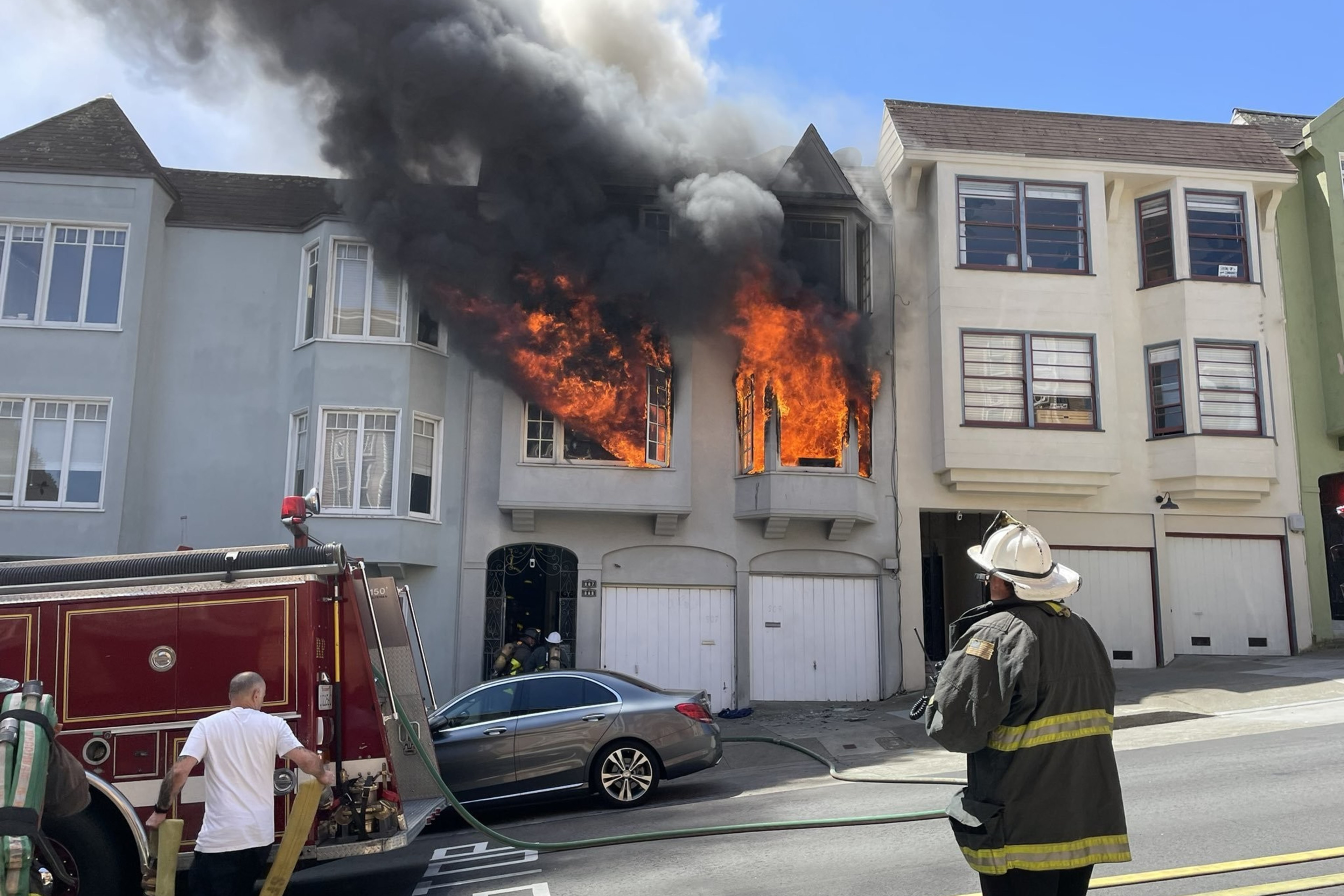 A building's upper floors are engulfed in flames, emitting thick black smoke, as firefighters, including one observing, and a fire truck are present.