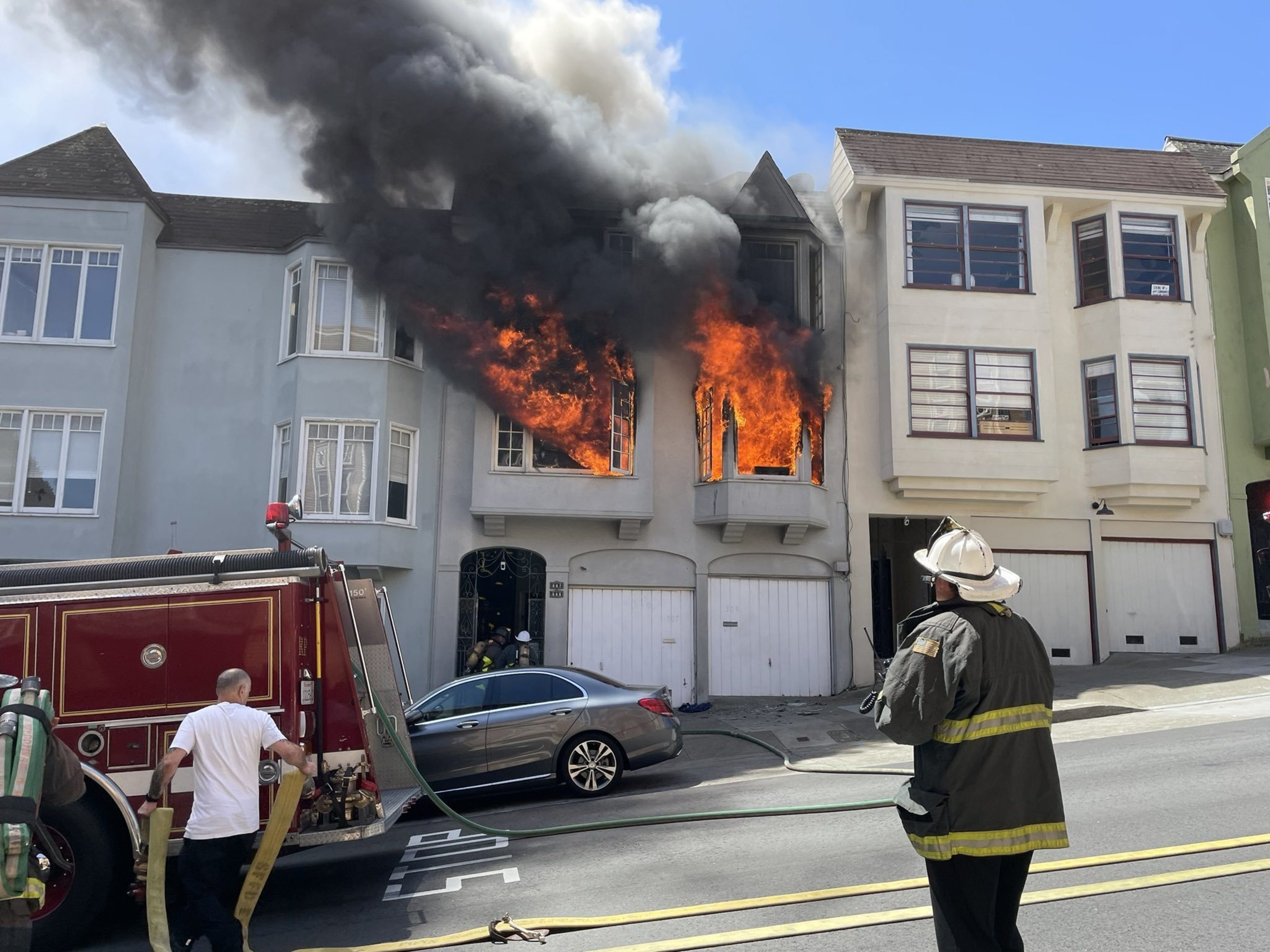 A building's upper floors are engulfed in flames, emitting thick black smoke, as firefighters, including one observing, and a fire truck are present.
