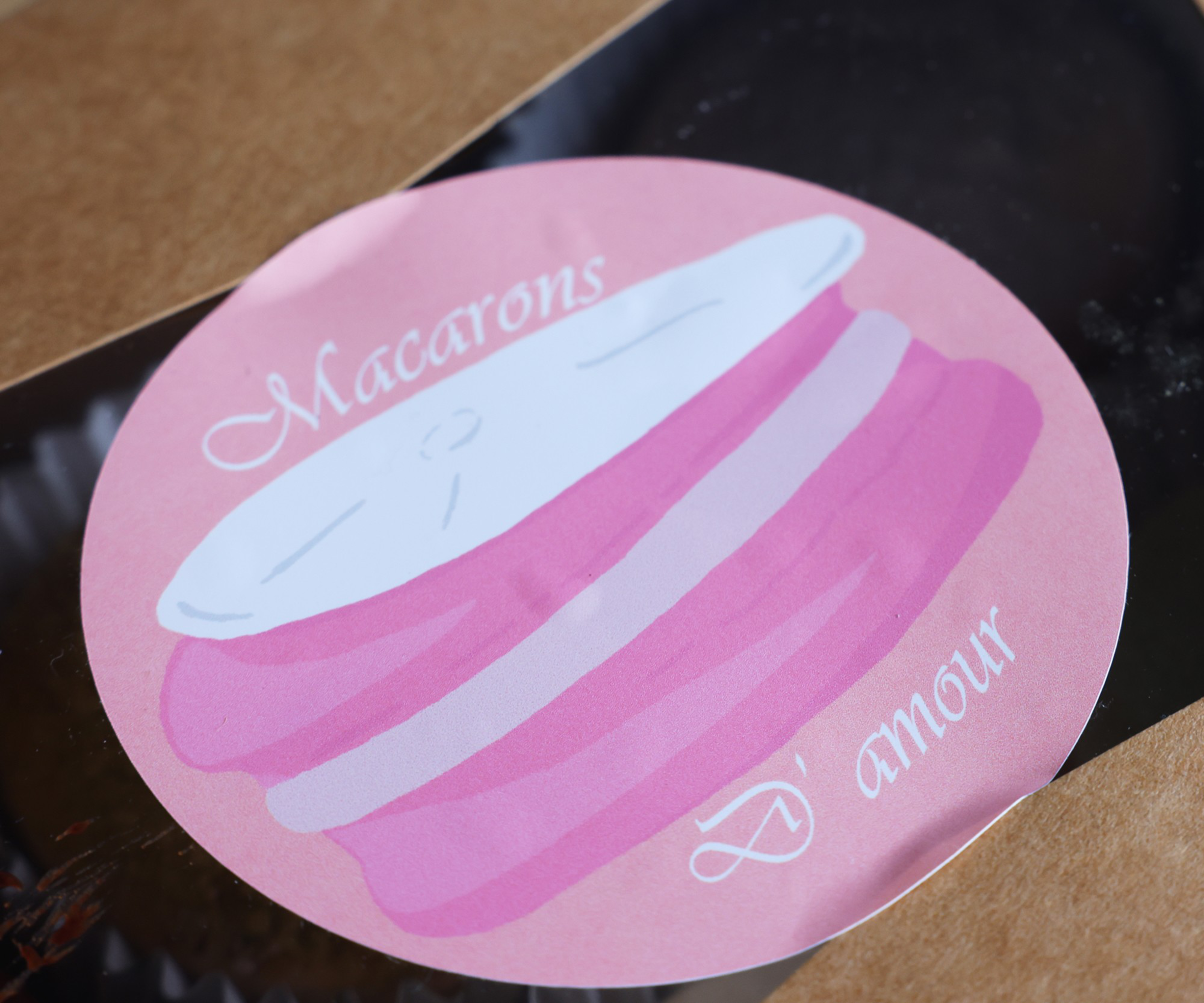 A round pink sticker on a box features an illustration of a pink and white macaron with the words "Macarons D' amour" written in white cursive.