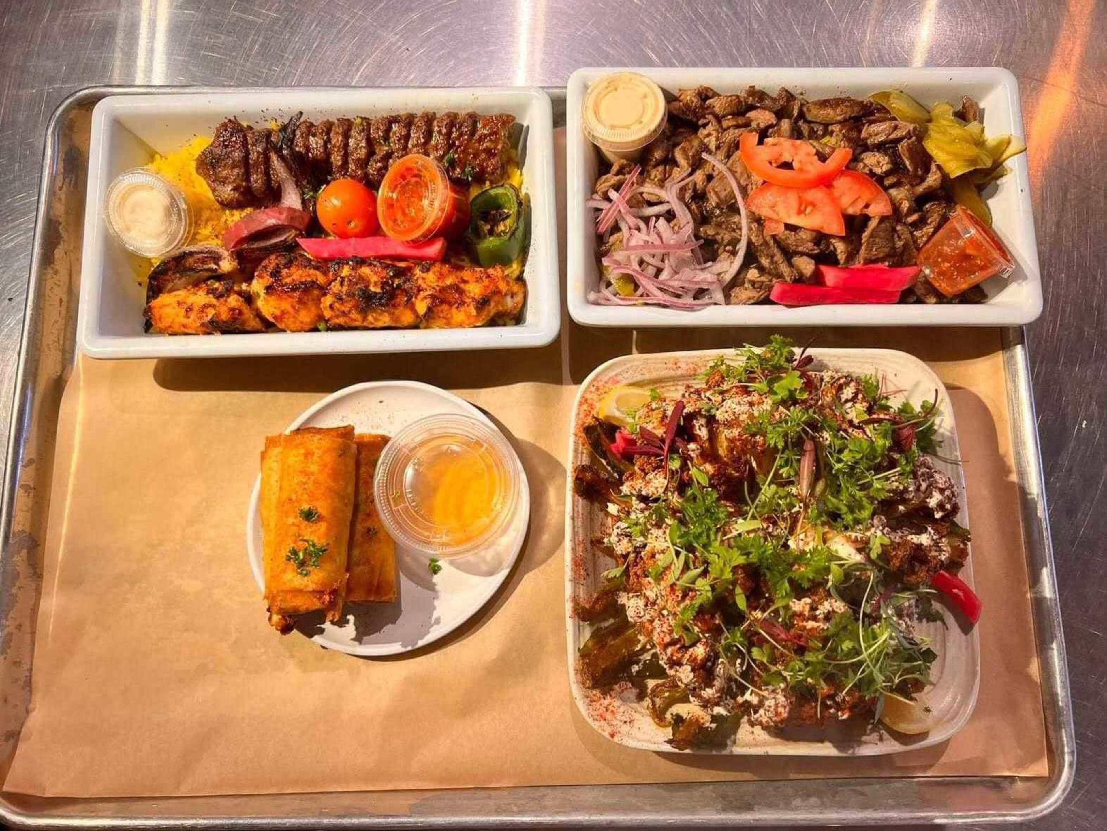 Two rectangular trays with grilled meat, vegetables, and sauces; a plate with two spring rolls and a dipping sauce; and a square plate of a salad with herbs and creamy dressing.