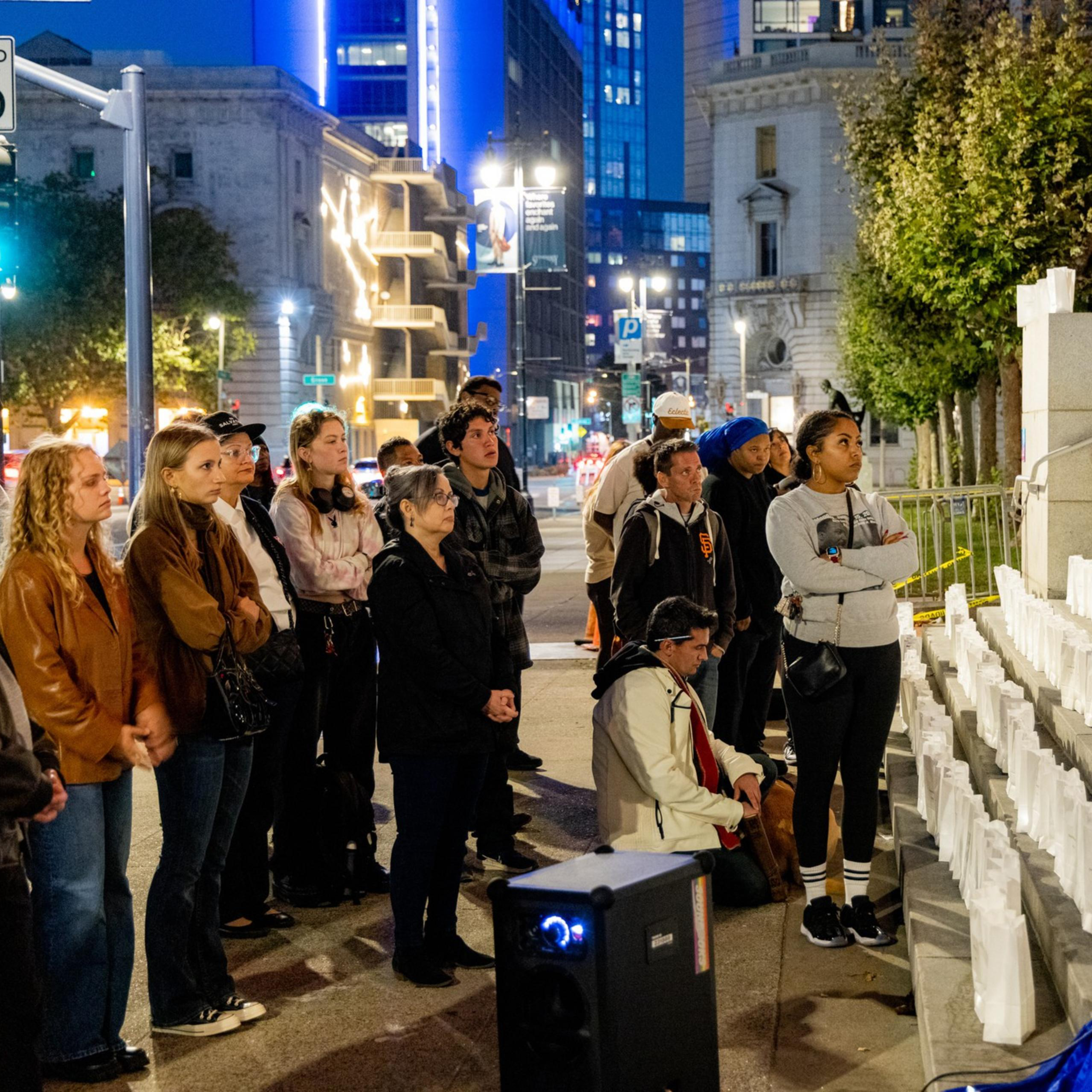 A group of people stands solemnly in an urban setting at night, facing a staircase lined with white paper lanterns. The city lights and buildings illuminate the background.