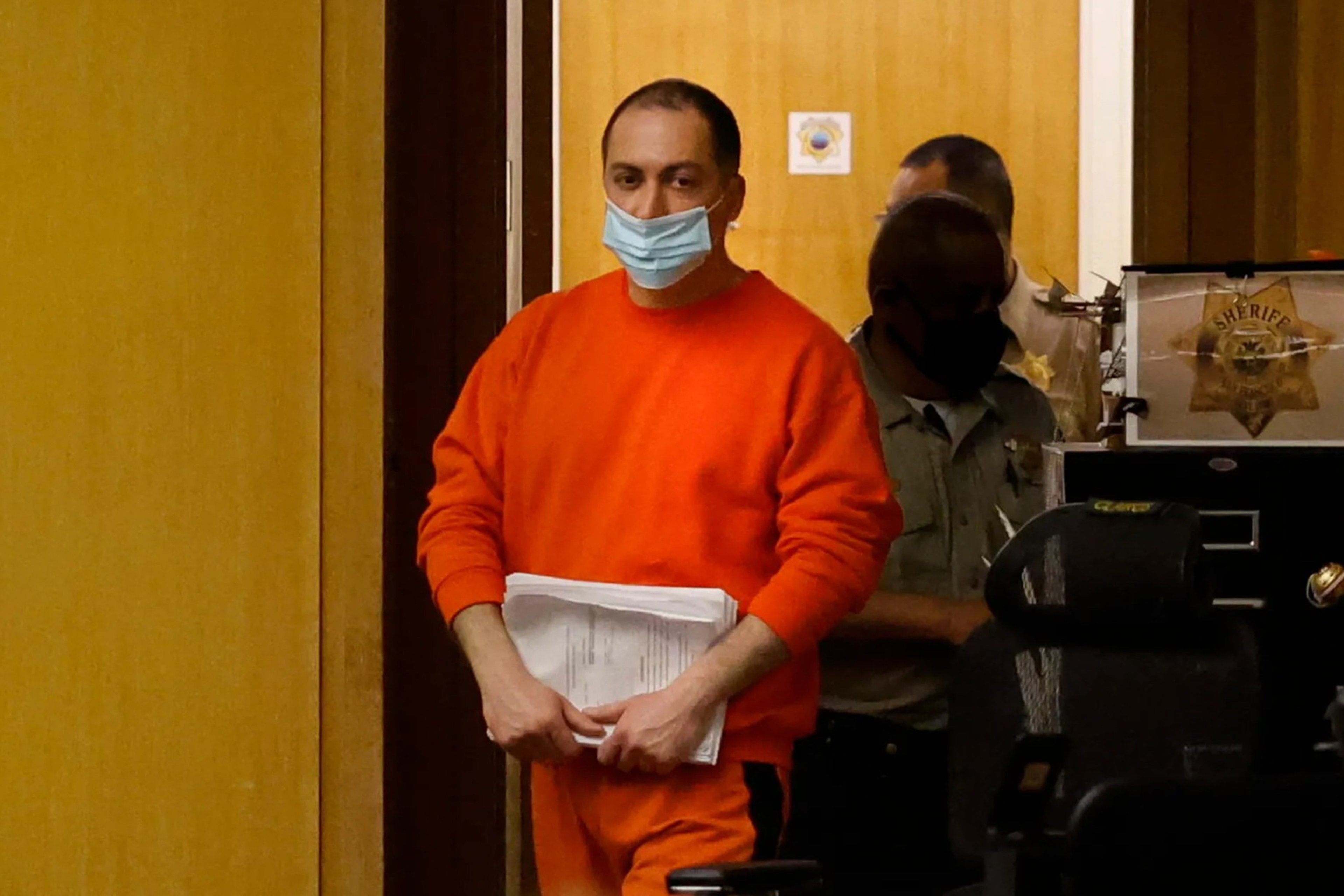 A person wearing an orange jumpsuit and blue mask stands in a courtroom holding papers. A sheriff's officer is visible behind them.