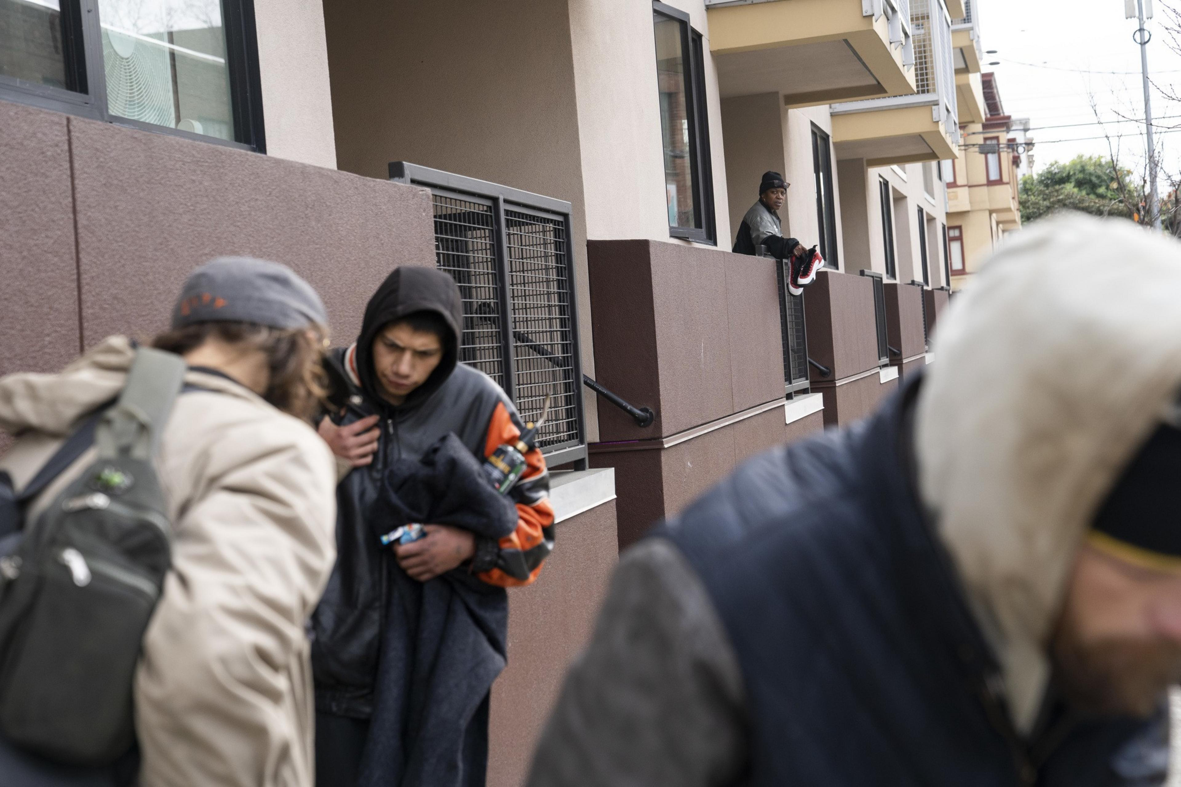 Three people in jackets walk by a building with maroon walls and windows. Another person is leaning on a balcony, holding shoes. The scene feels urban.