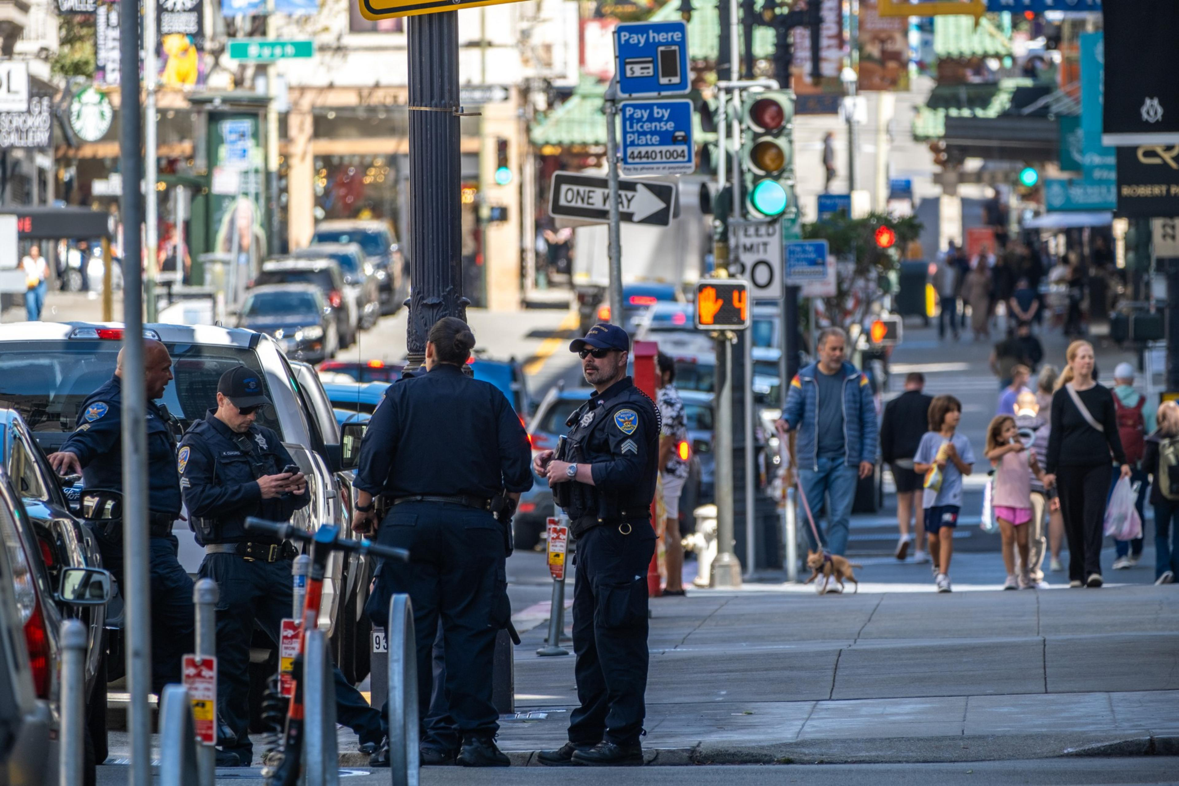 The image shows several uniformed police officers standing and talking on a busy urban street, with pedestrians, street signs, and buildings visible in the background.