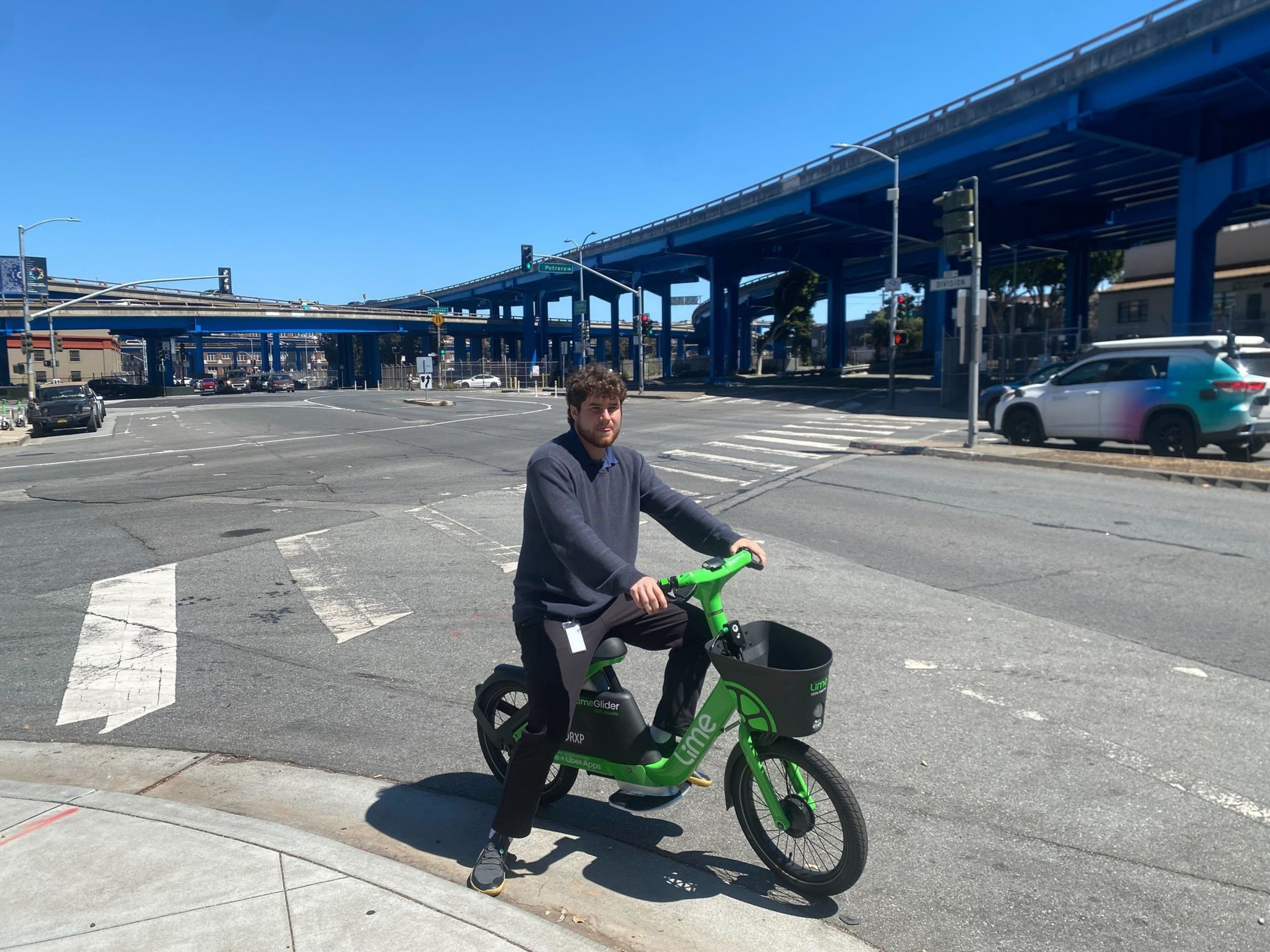 A man wearing a dark sweater and pants sits on a green Lime electric bike at a street corner under blue highway overpasses on a sunny day.