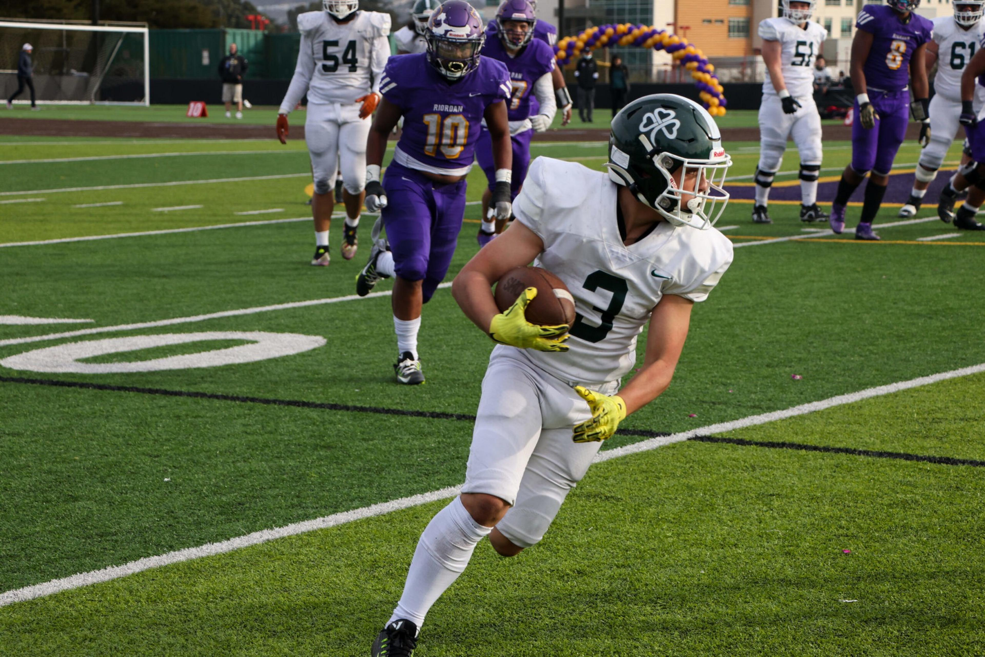 A football player in a white uniform and green helmet runs with the ball near the sideline as players in purple and white uniforms chase him.