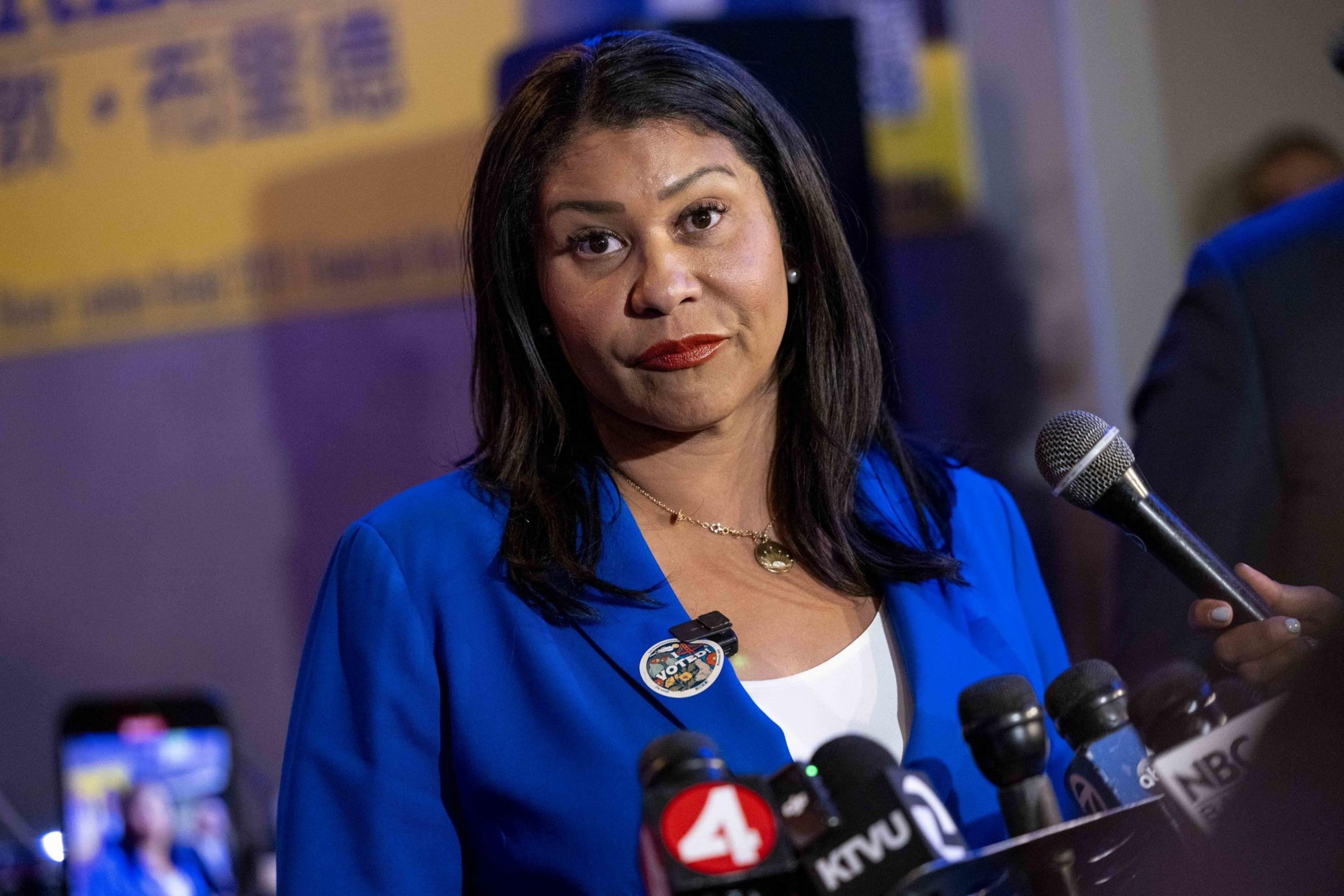 A woman in a blue blazer stands in front of multiple microphones, speaking at a press event. She is wearing a pin that says &quot;VOTE&quot; and appears focused and composed.