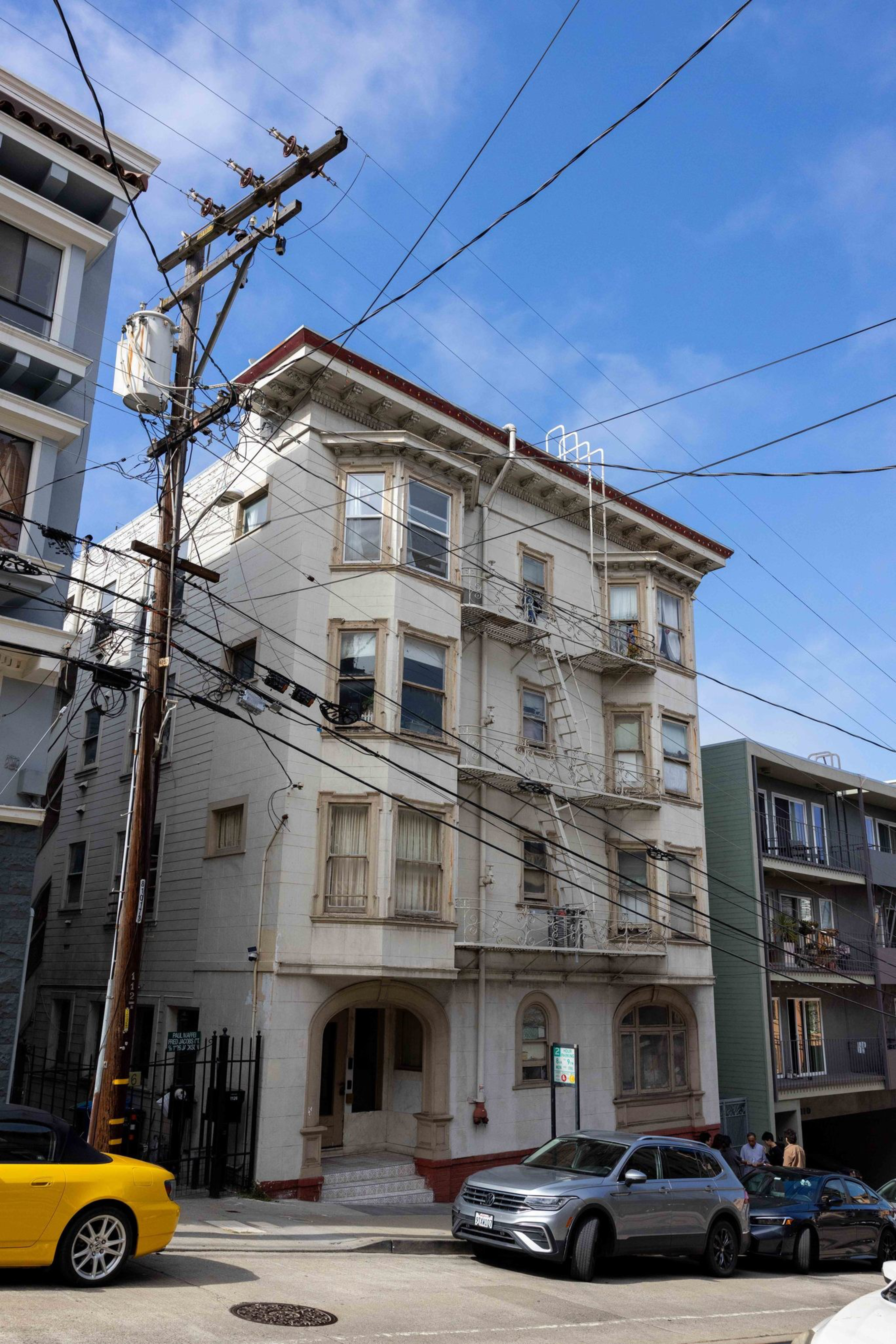 A four-story beige building with fire escapes, multiple windows, and cars parked along a steep street under a clear blue sky.