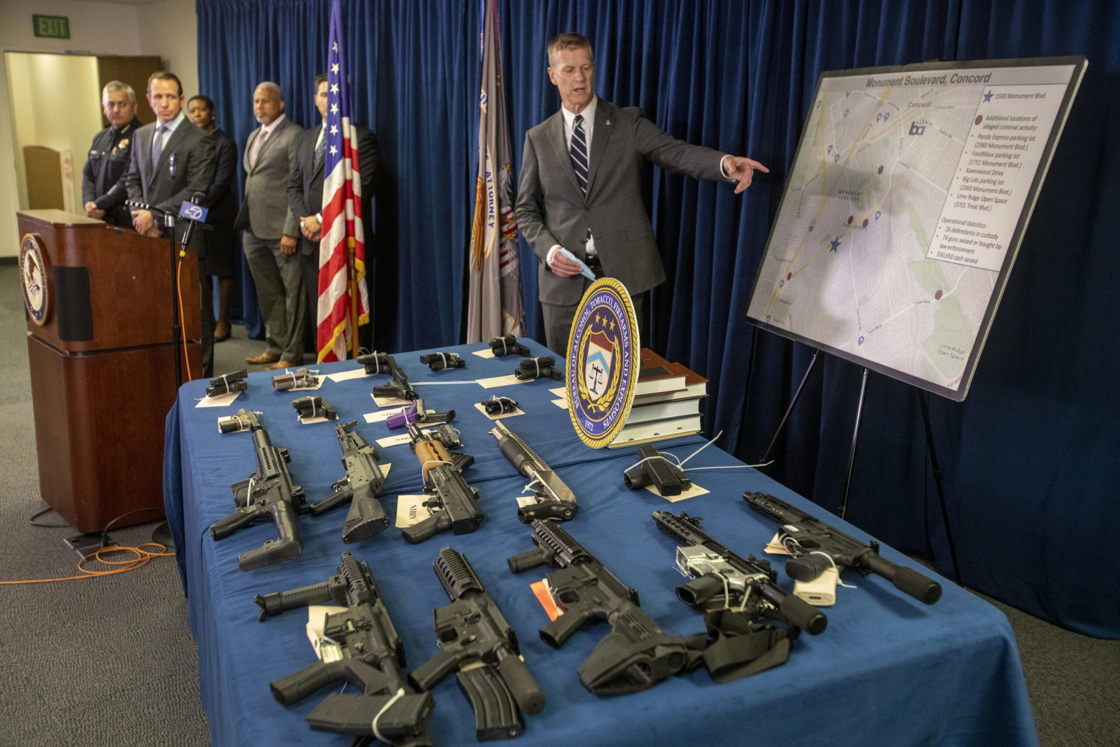 A man in a suit points to a map on an easel while other officials stand near a podium; multiple firearms are displayed on a blue-covered table.