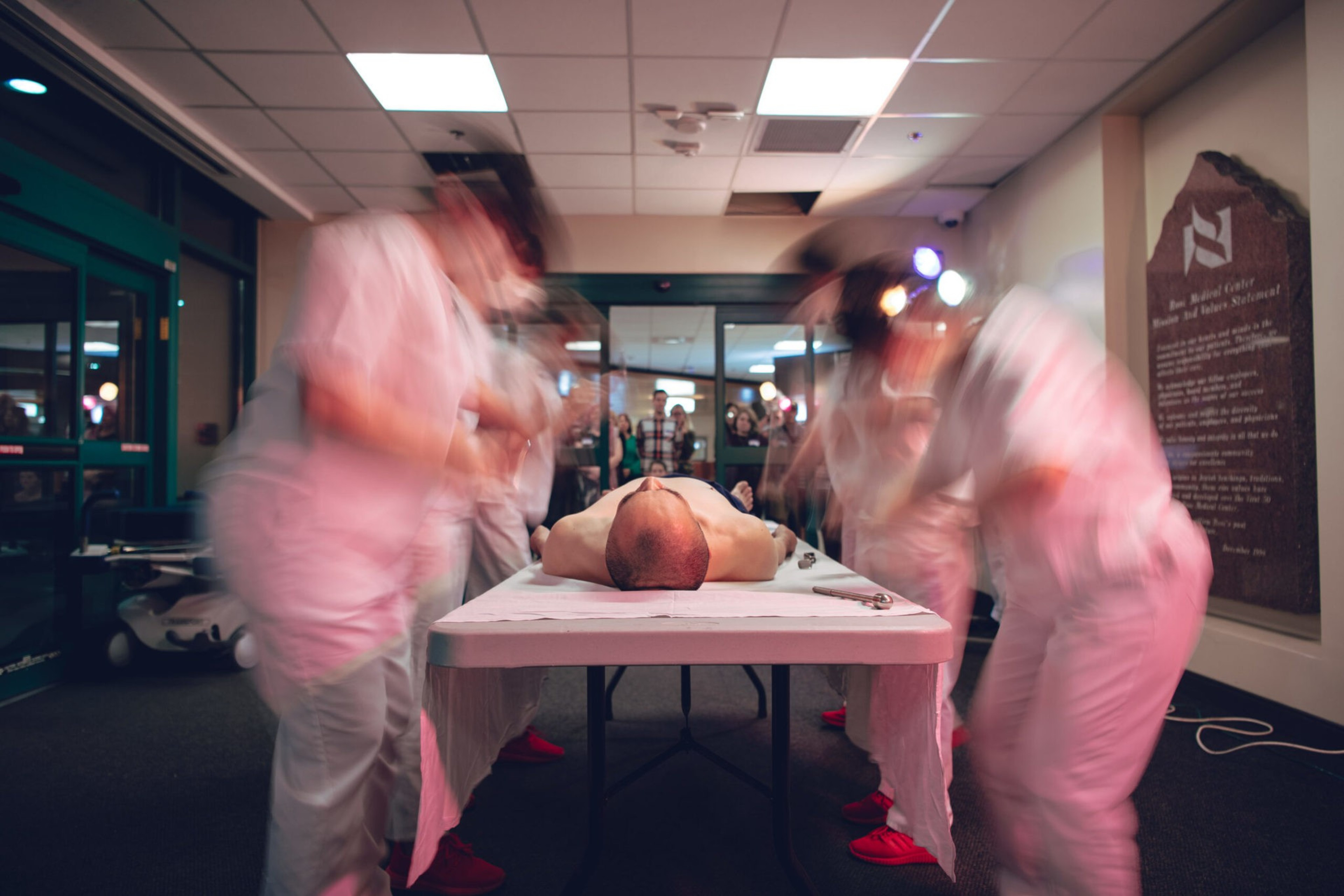 A man lies motionless on a table as three blurred figures in white uniforms rush around him in a brightly lit room with onlookers in the background.