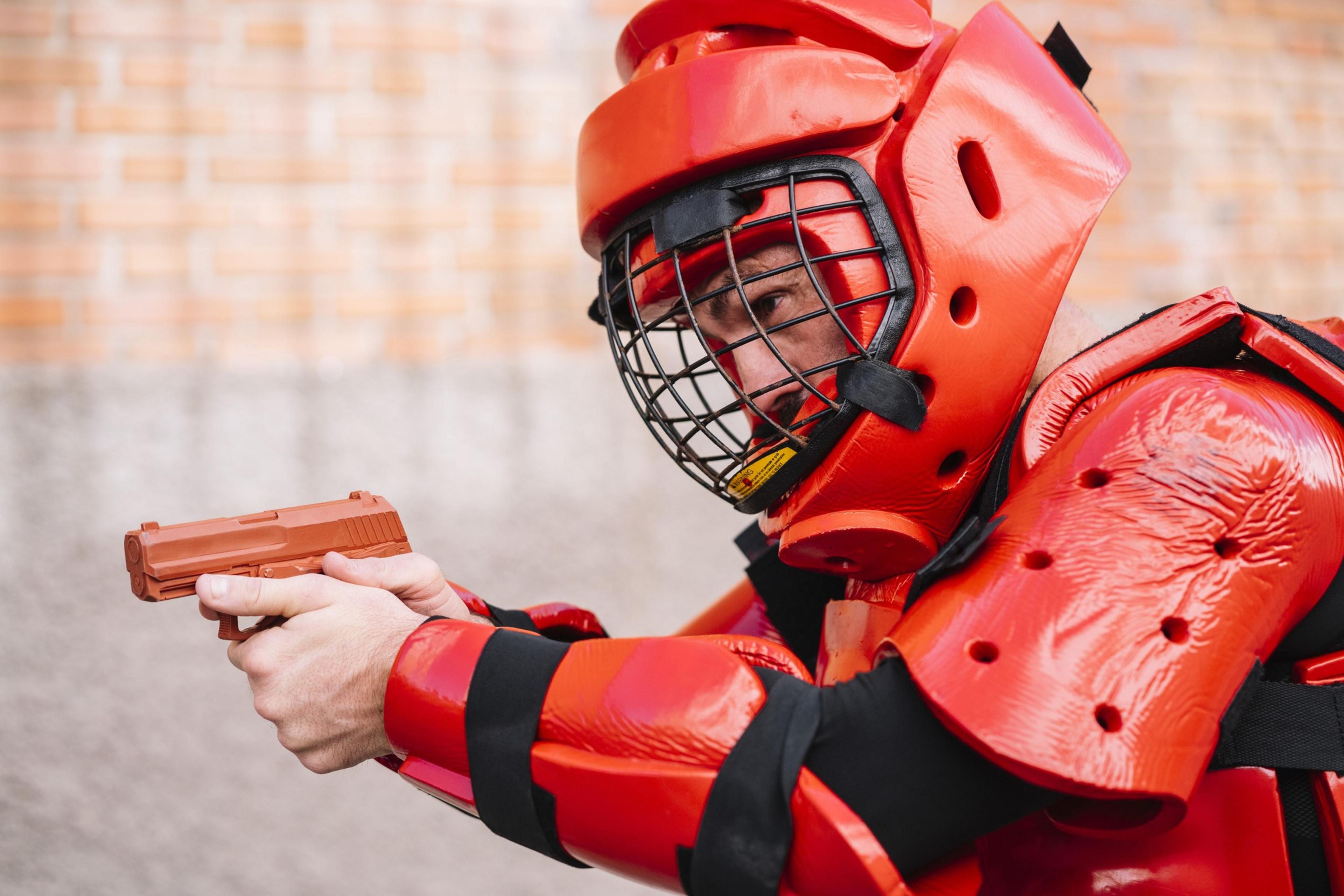 A person wearing bright red protective gear and a helmet with a face guard is aiming a brown practice gun.