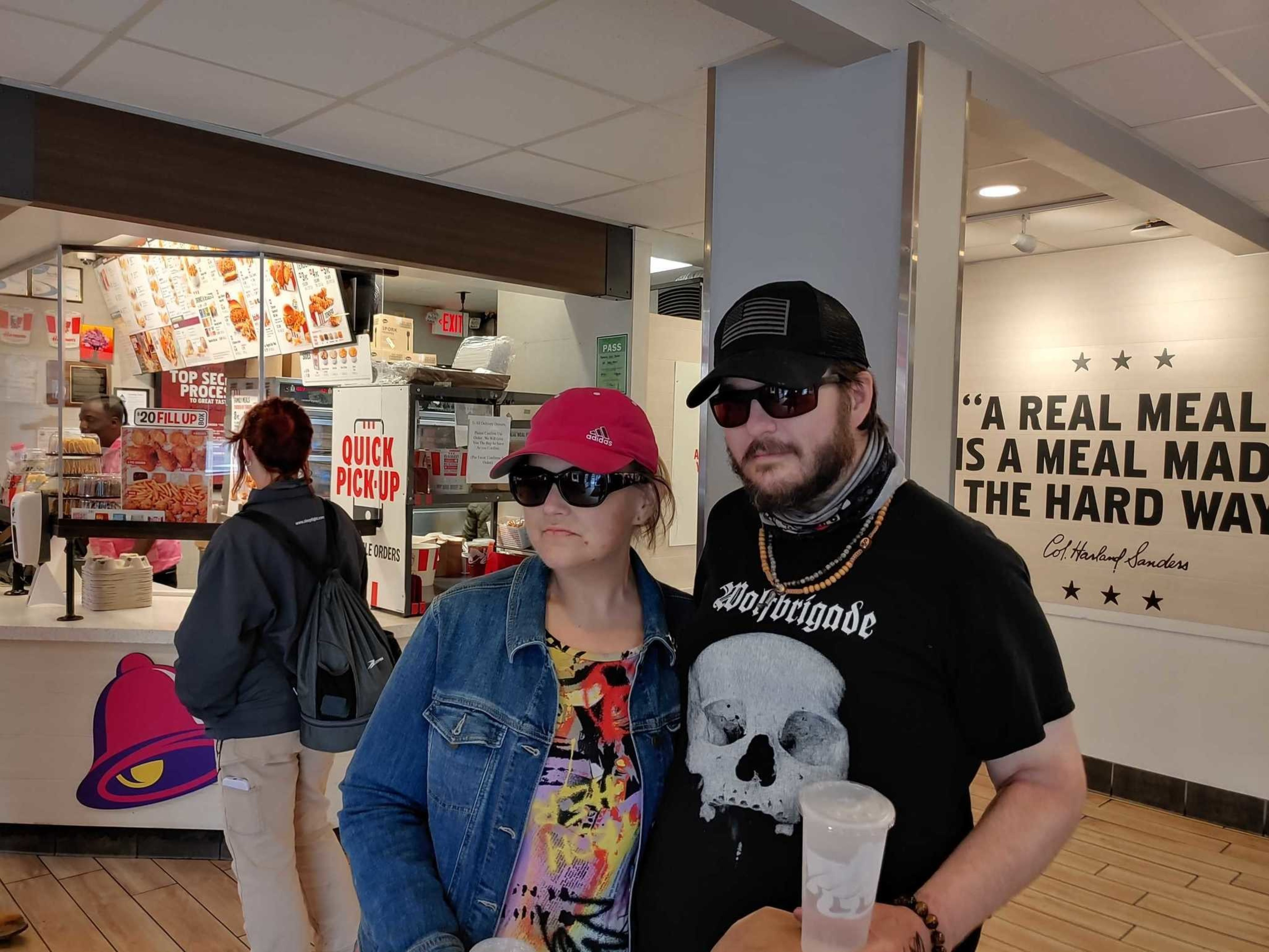 A man and woman wearing sunglasses and casual clothes stand inside a fast-food restaurant with a "Quick Pick Up" sign and a Colonel Sanders quote on the wall.
