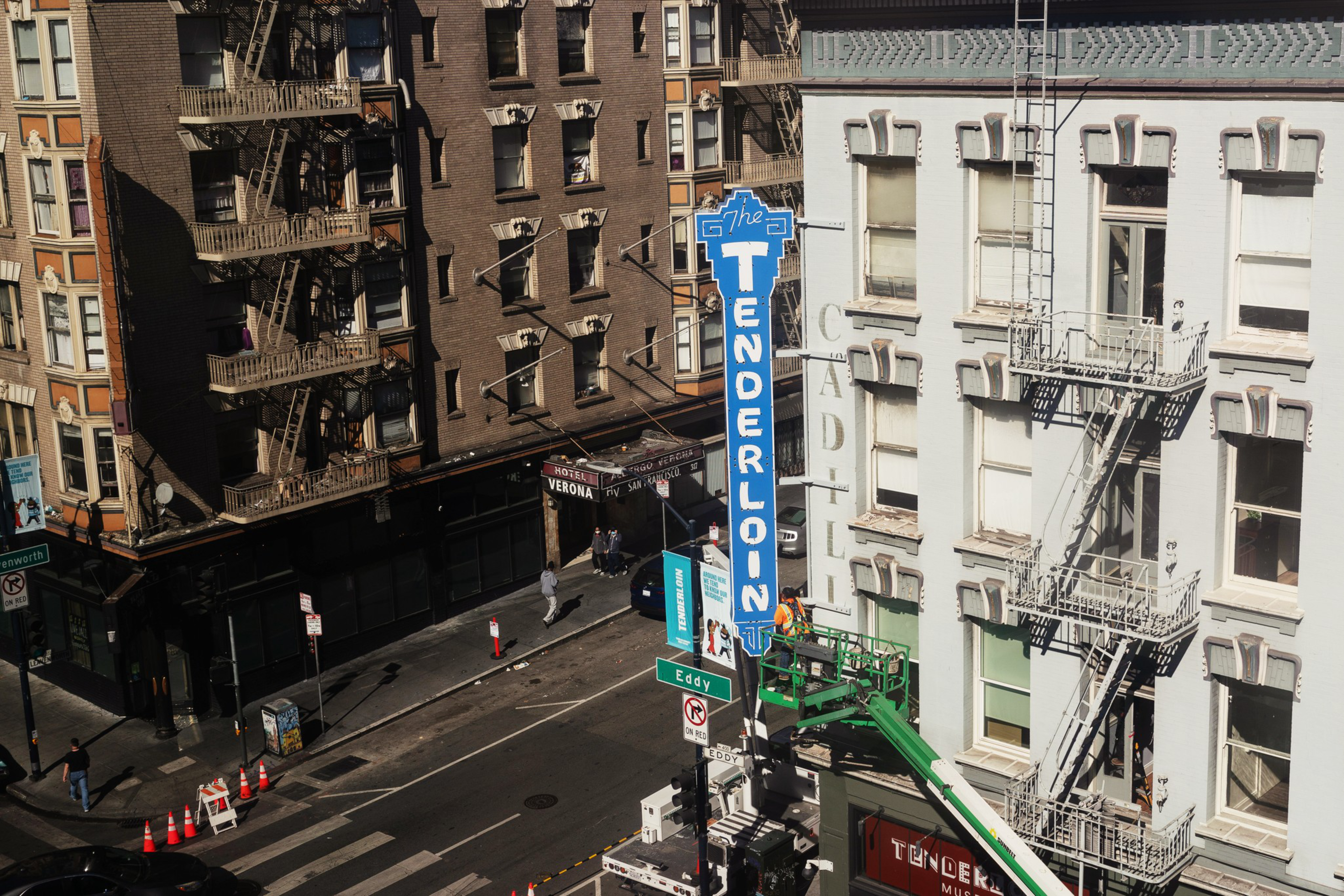 A city street corner with tall buildings, fire escapes, and a blue "The Tenderloin" sign.