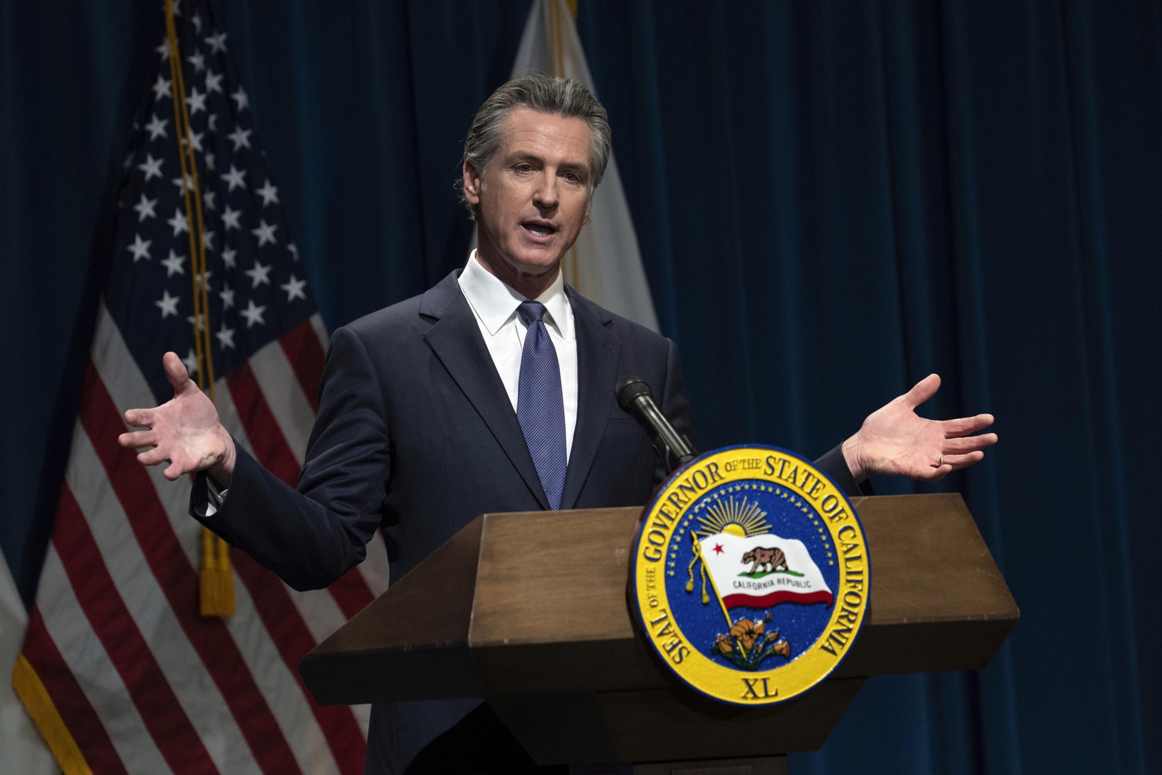A man is speaking at a podium with the California state seal, flanked by US flags.